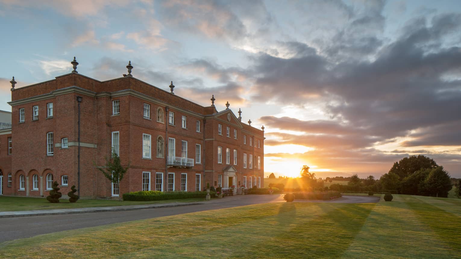 A large red brick building next to a road and lush grass with the sun setting behind it.