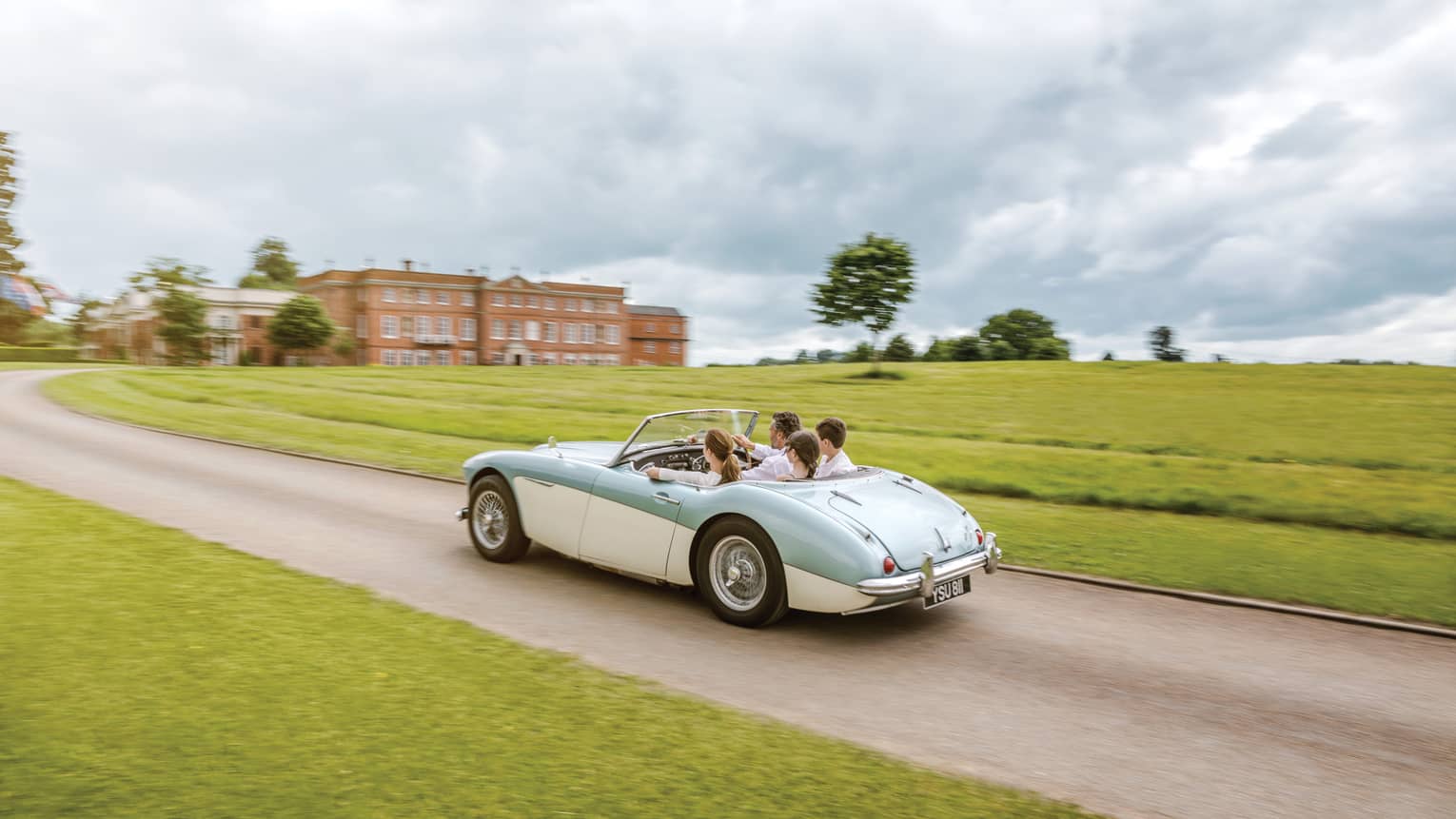 Family rides in vintage convertible car down road past English countryside to hotel manor