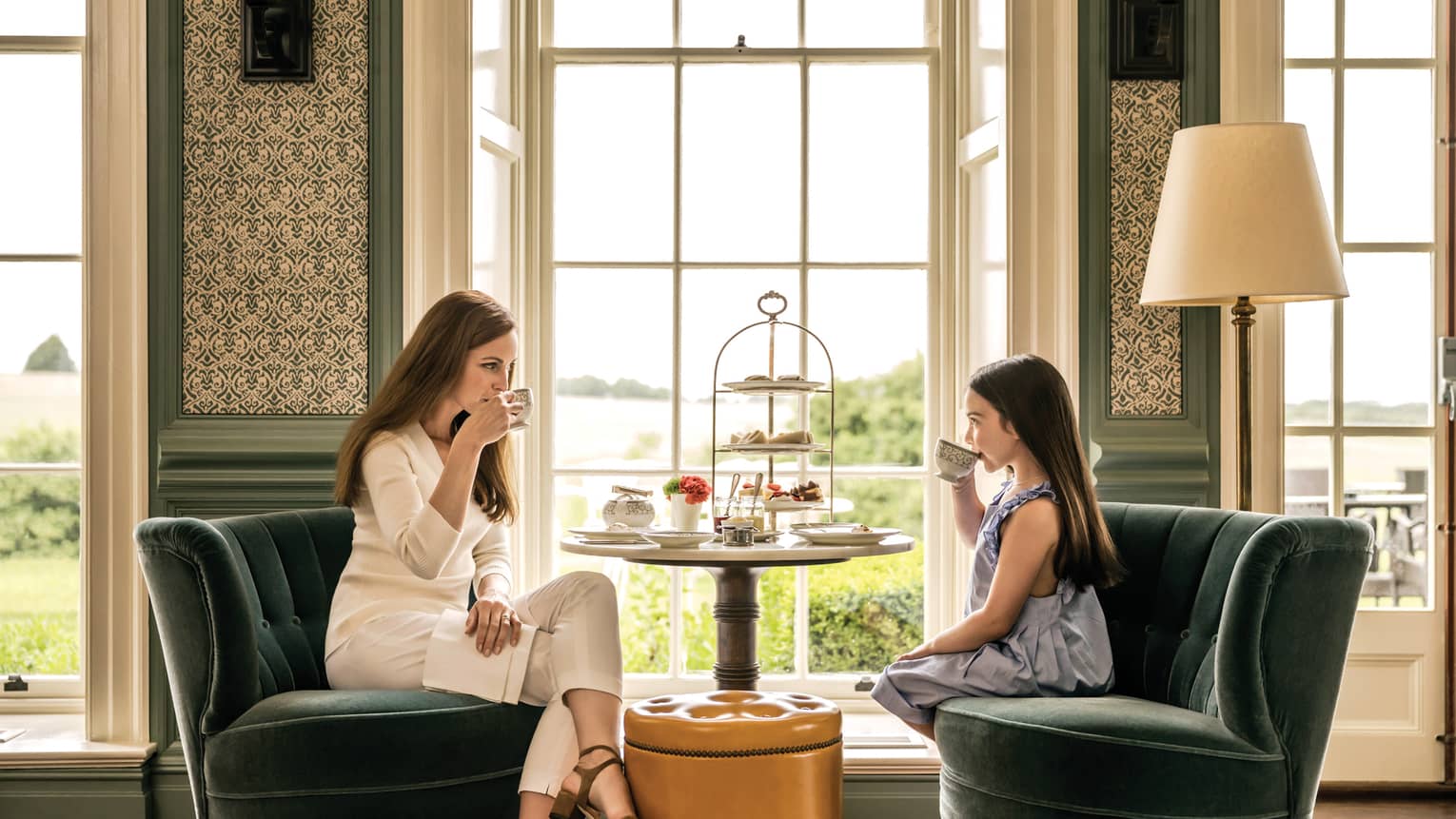 Woman and young girl sit in velvet armchairs by sunny window, enjoy afternoon tea service