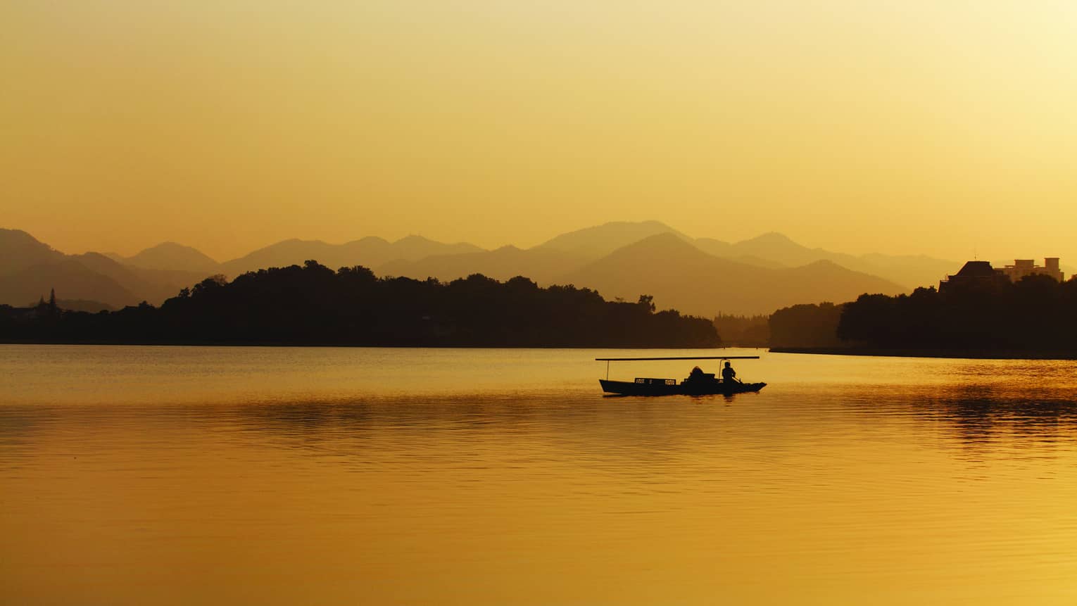 Long view of a lake under hazy golden glow, a small wooden boat silhouetted against treed islands and and undulating hills.
