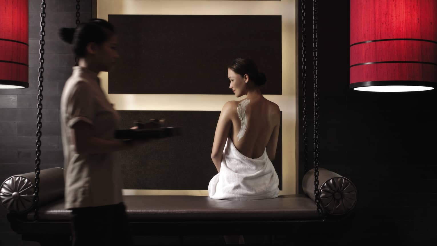 Woman in white towel with cream on back sits on bench in dark spa room