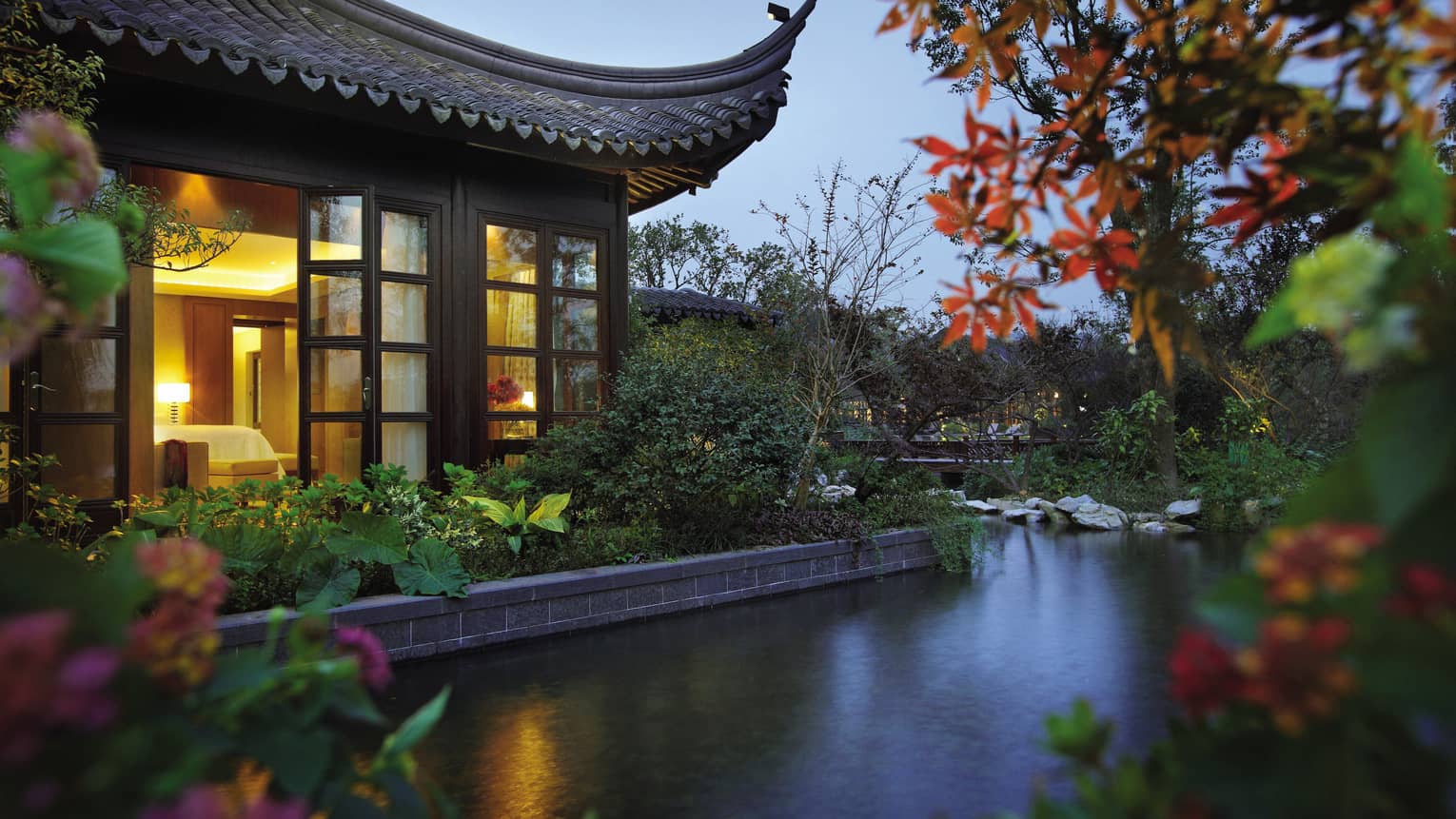 Side view of Lake Suite villa with sweeping roof, garden over water at night 
