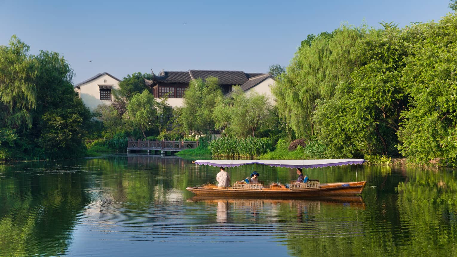 Three people sit in a small wooden boat with a flat canopy, surrounded by ripples in the water near a shoreline with trees.