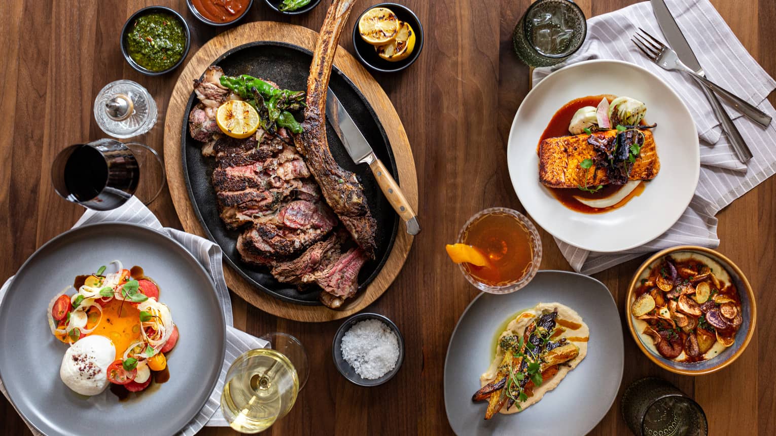 An assortment of food including fish, steak, salad, and different sauces on grey and white plates.