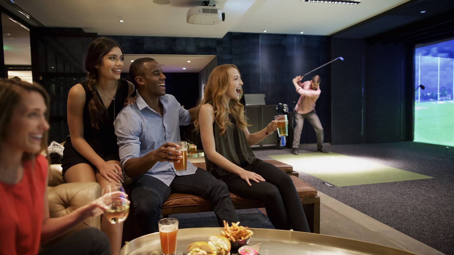 A man and three woman sip beer and cheer on a sports team in the foreground, while another man golfs in the background of a function room at four seasons hotel houston