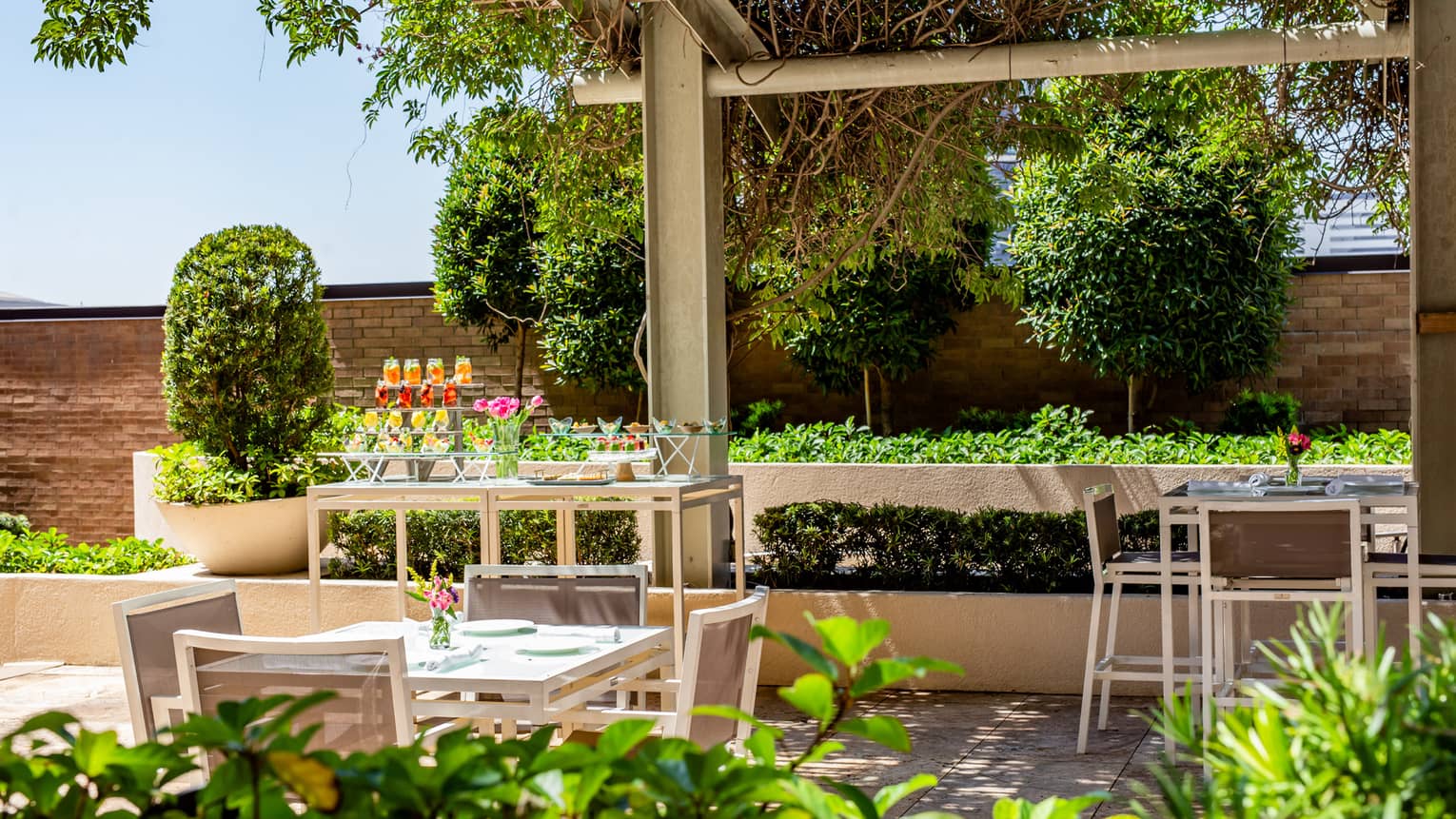 An outdoor event space with square tables and chairs surrounded by green plants.