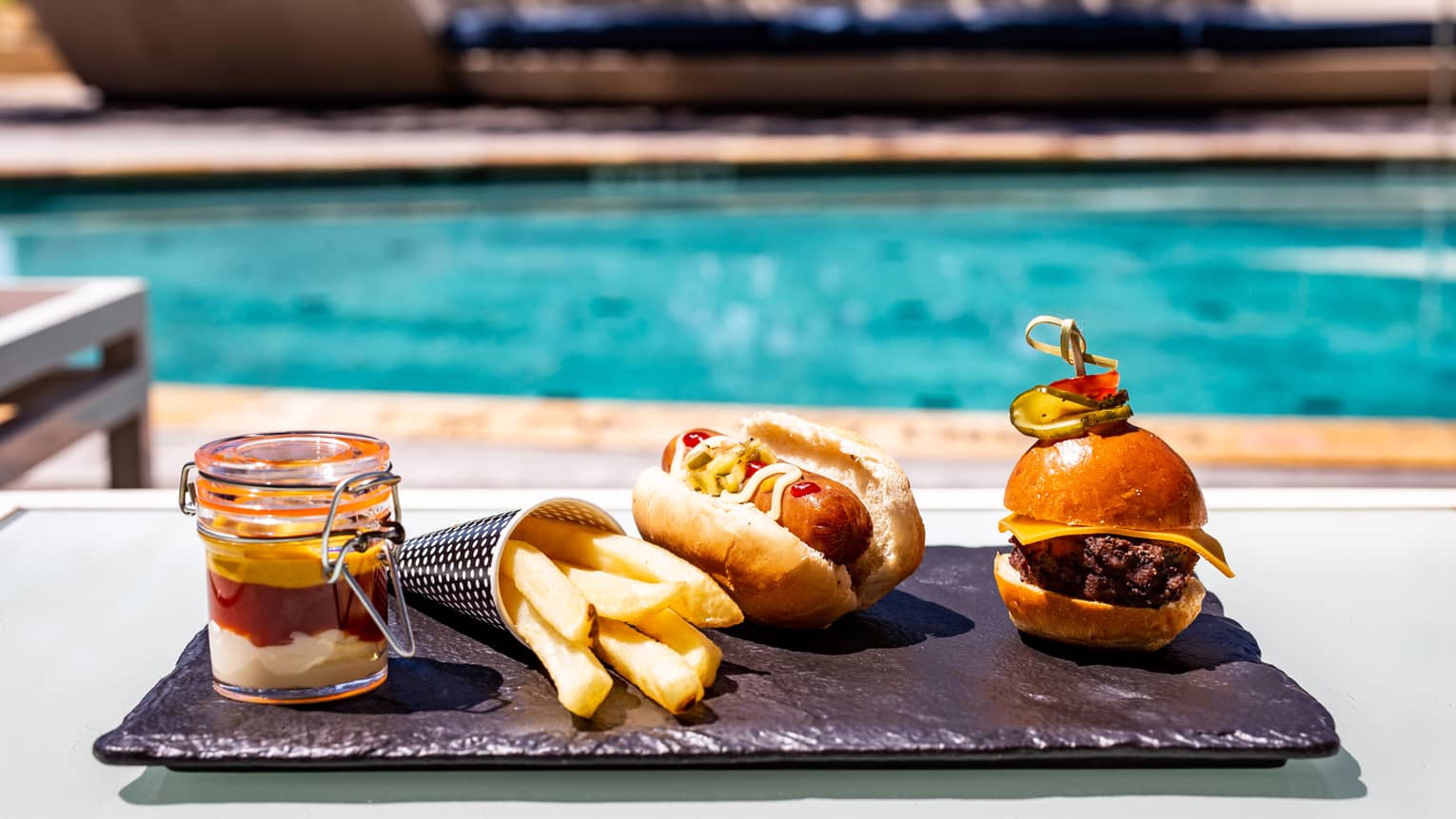 A burger, hotdog, fries, and drink on a dark tray near a pool.