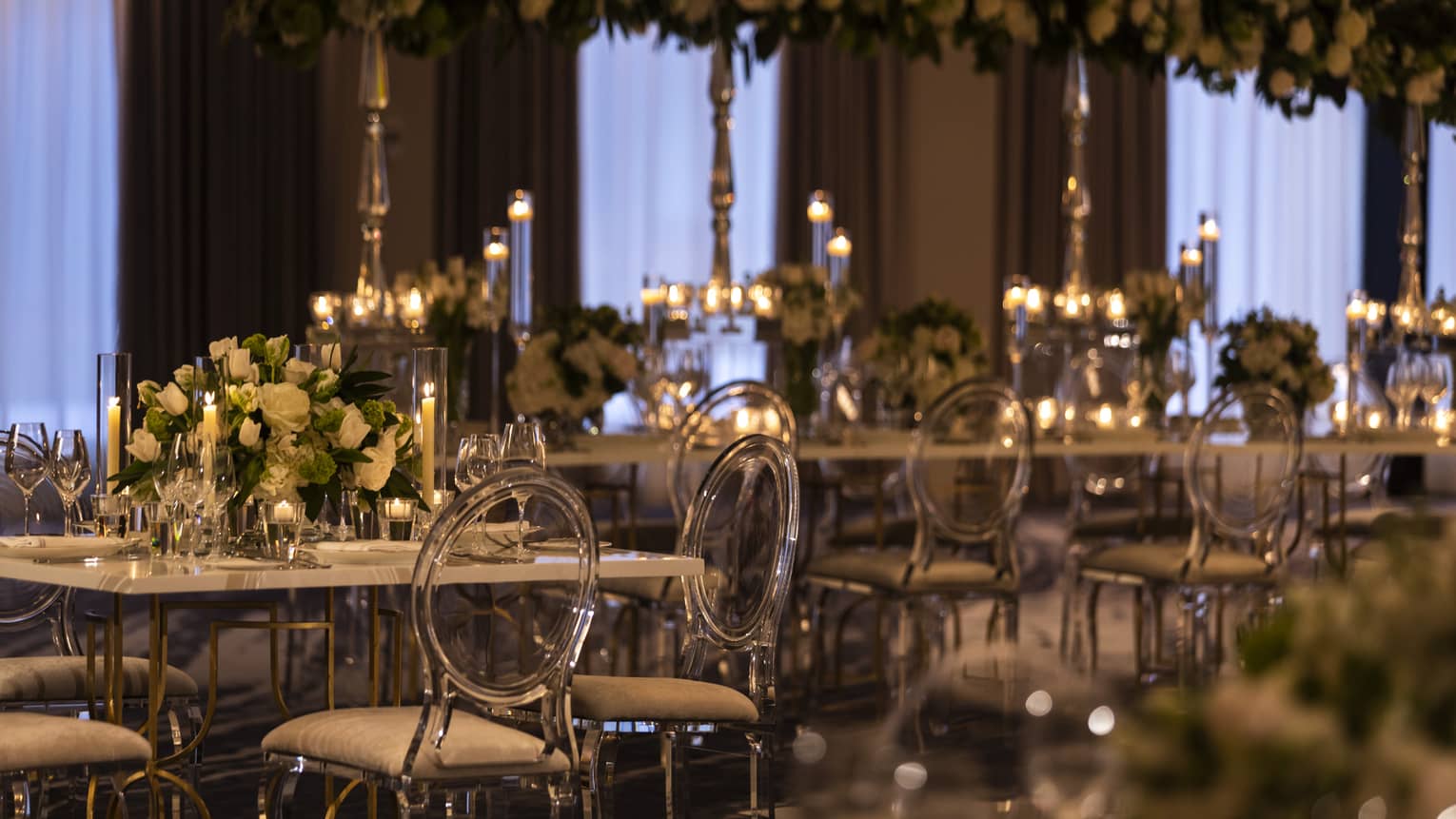 A dimly lit ball room with long tables and clear chairs is covered in flowers and candles.