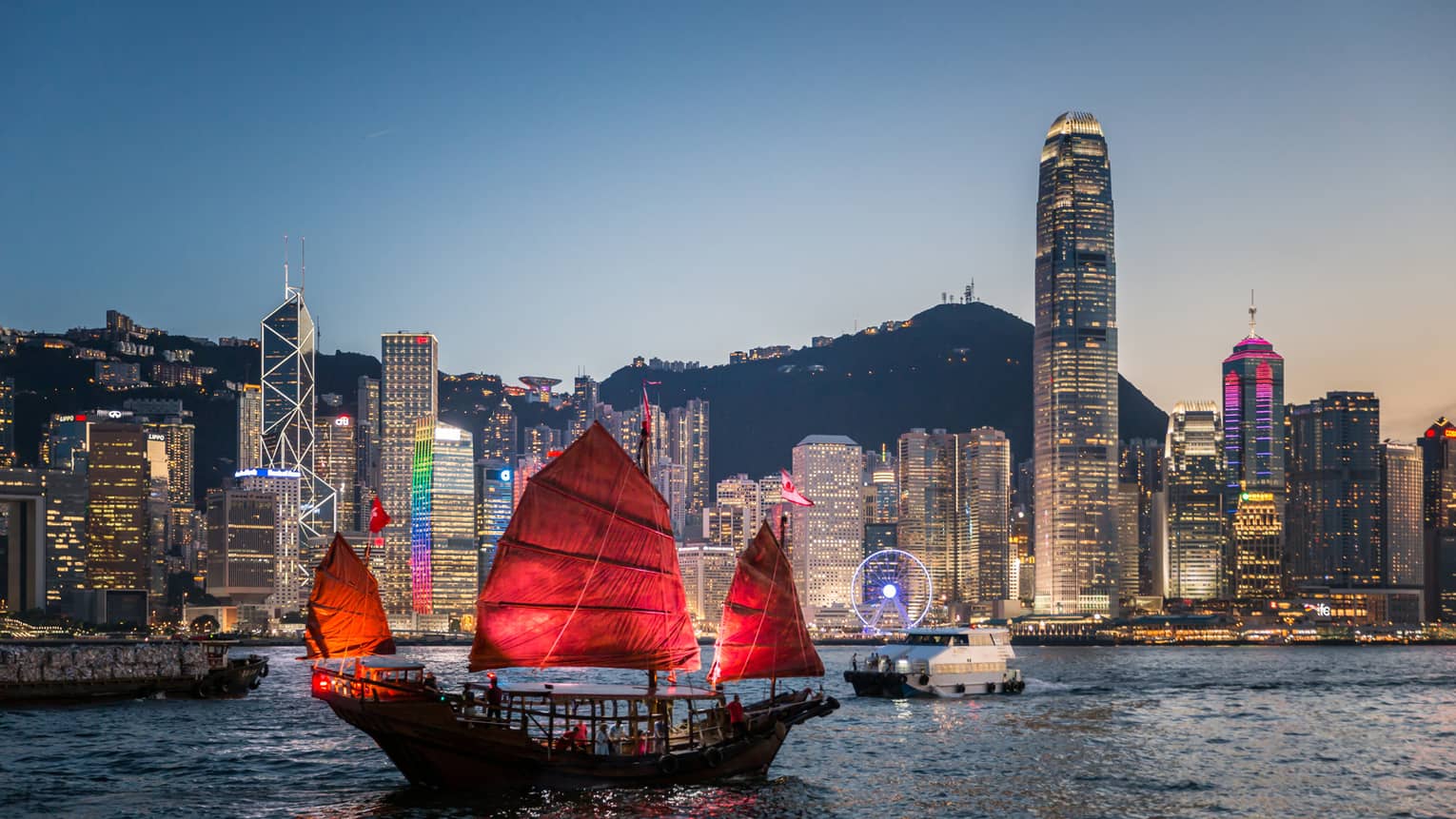 A traditional Hong Kong junk boat with red sails gliding through Victoria Harbour at dusk, with the city’s illuminated skyline in the background