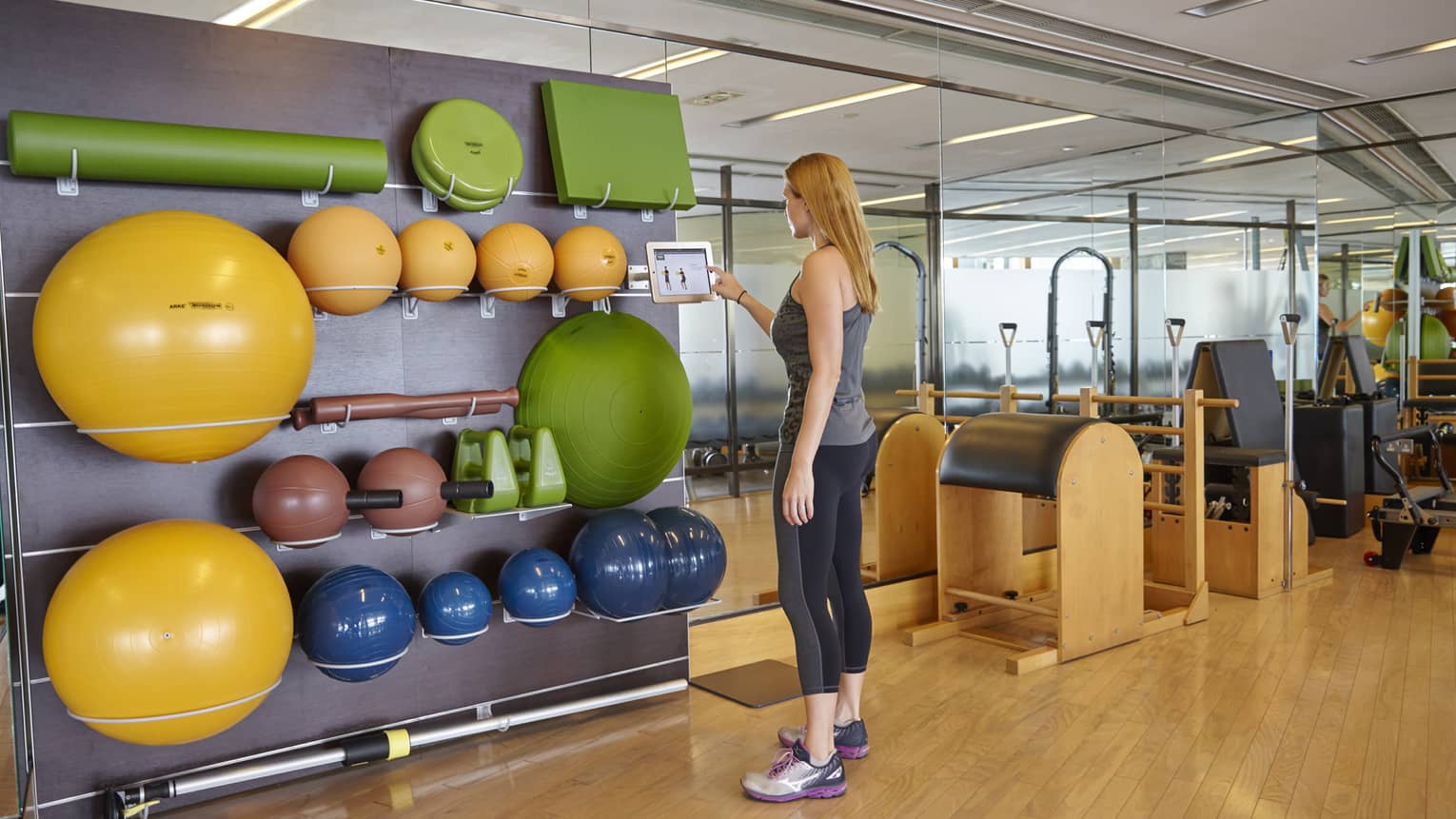 A person in athletic wear stands in a modern gym, interacting with a tablet mounted on a wall. Surrounding her are various exercise balls, mats and equipment.