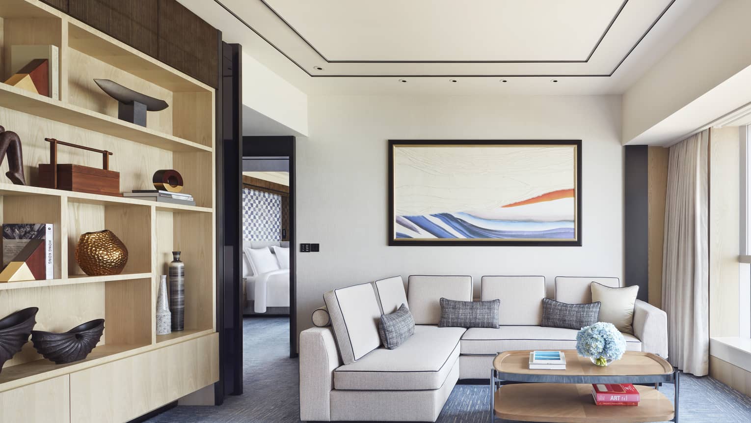 Modern hotel living area with beige sectional sofa, abstract wall art, open shelving with decor and coffee table with books and blue flowers.