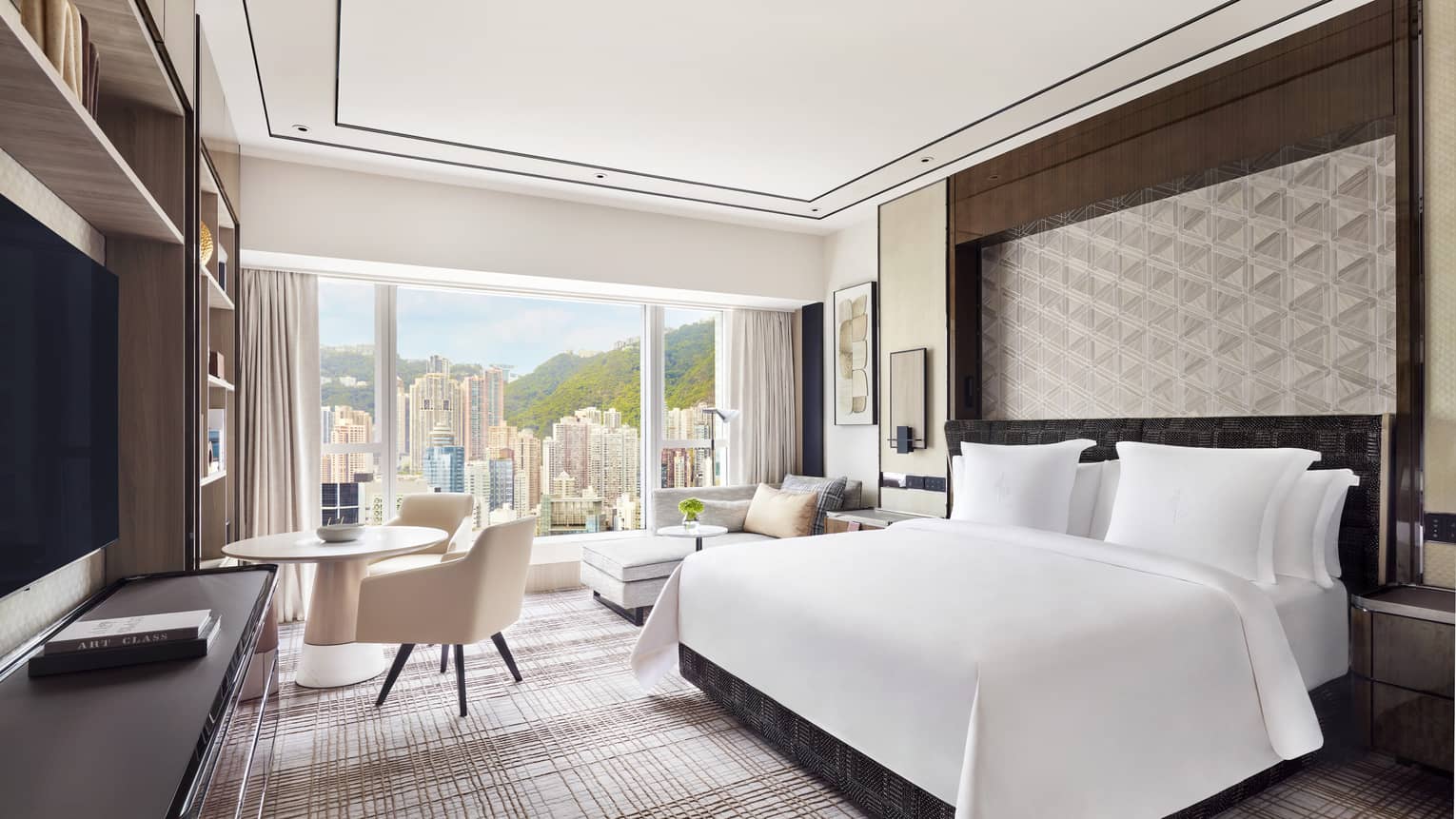 Modern hotel room with a large bed, geometric wall design, and a seating area next to a round table. Floor-to-ceiling windows offer a view of skyscrapers.