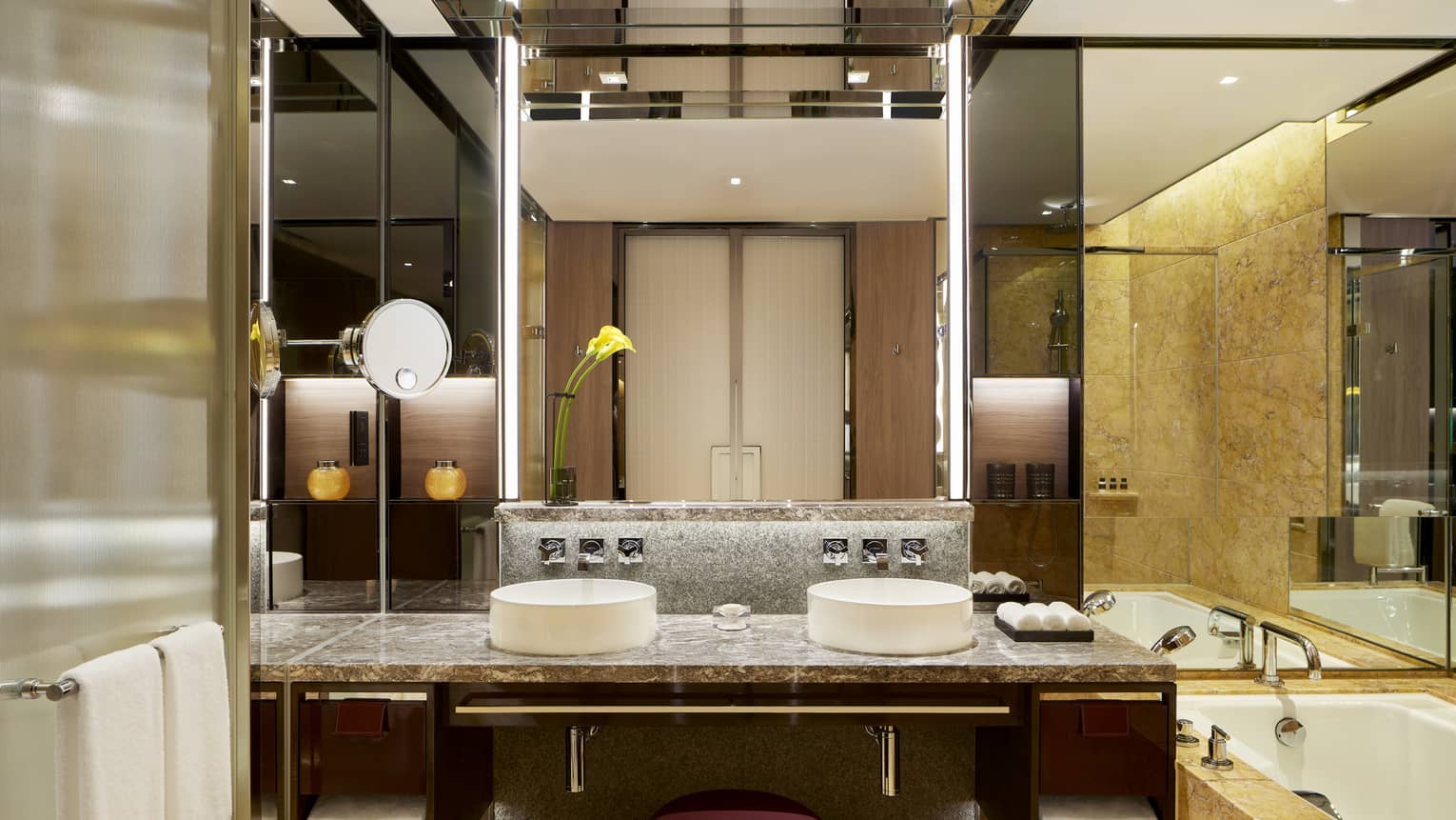 Modern bathroom with double sinks, marble countertops, a large mirror with backlighting, and a bathtub. A yellow flower in a vase decorates the counter.