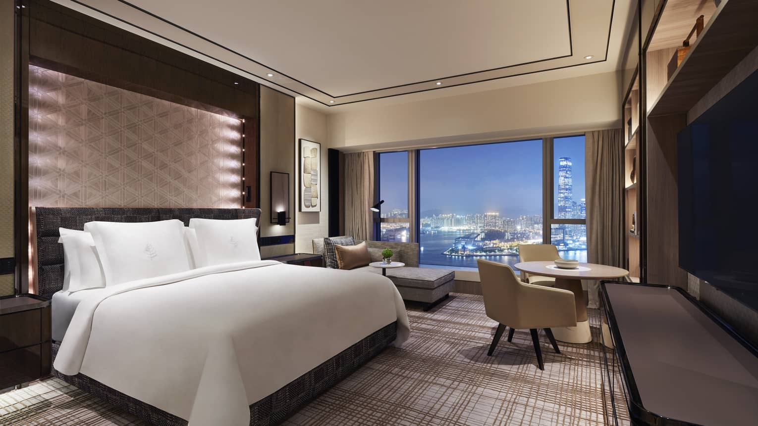Hotel room with king bed, plaid carpet, TV, chairs, view of city at night