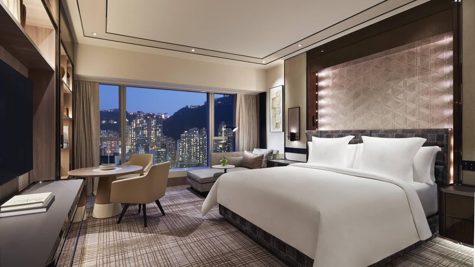 Luxury hotel room with a large bed, geometric accent wall, and a seating area. Floor-to-ceiling windows provide a nighttime view of city skyscrapers.