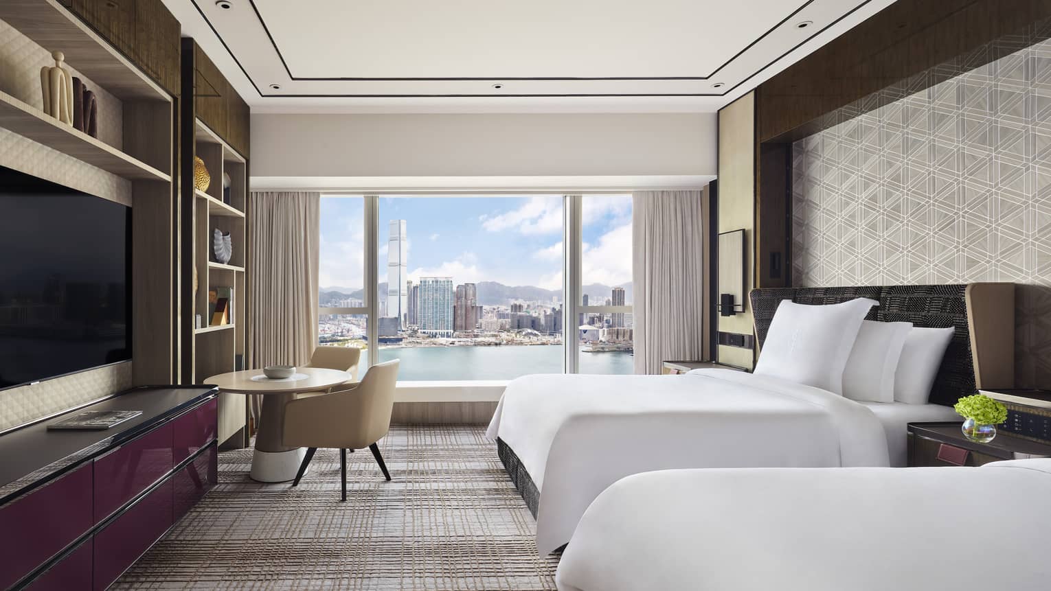Modern hotel room with two beds, and a round table by the window. Floor-to-ceiling windows offer a daytime view of the city skyline and waterfront.