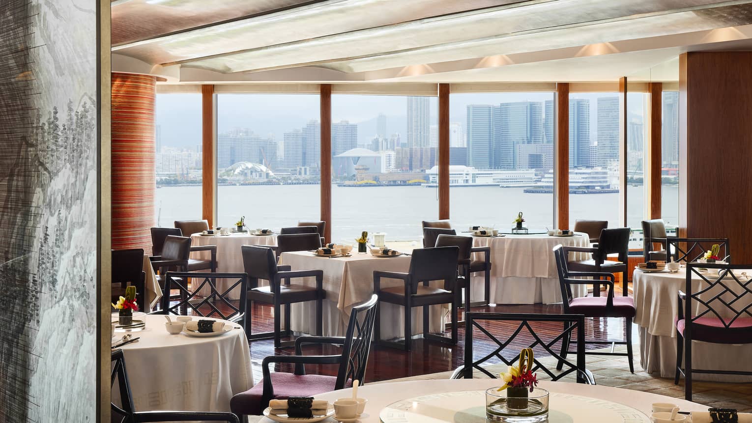 An elegant dining room with tables set with flowers and place settings, overlooking the city from across a body of water.