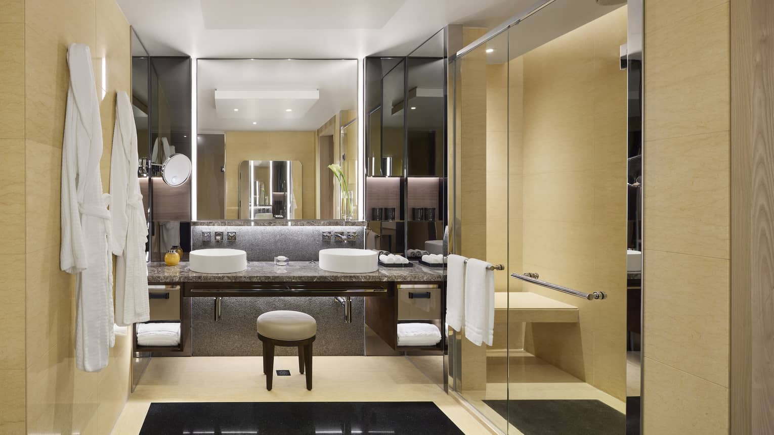 Luxurious hotel bathroom with double sinks, large mirrors, beige walls, glass shower and hanging white bathrobes.