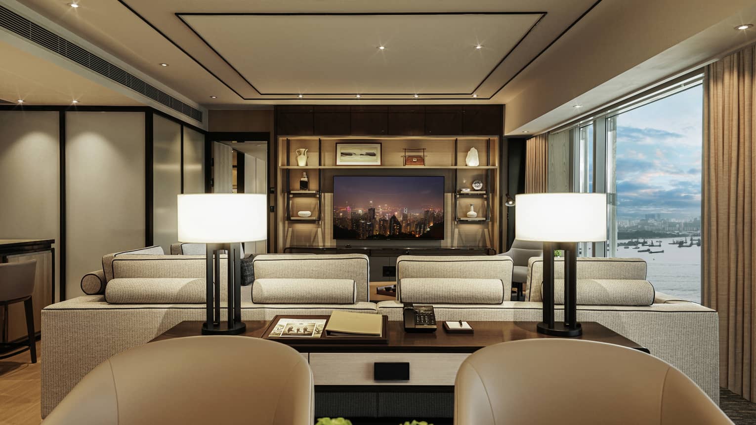 Luxurious hotel living room with large beige sofa, wall-mounted TV displaying city skyline, shelf decor and view of harbour through windows.
