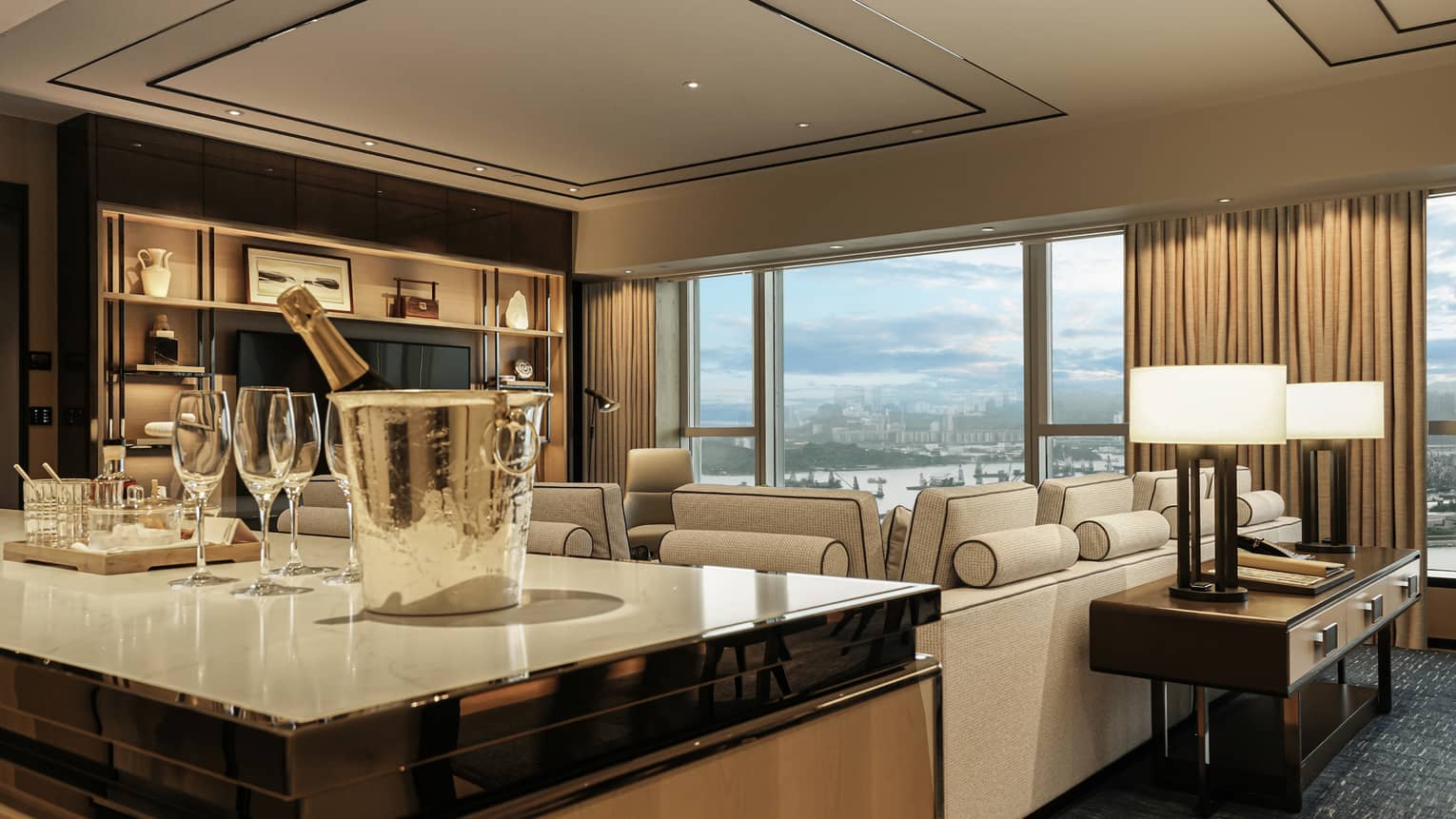 Luxurious hotel living room with champagne bucket, wine glasses, beige sectional sofa, large windows with city view and decorative shelving.