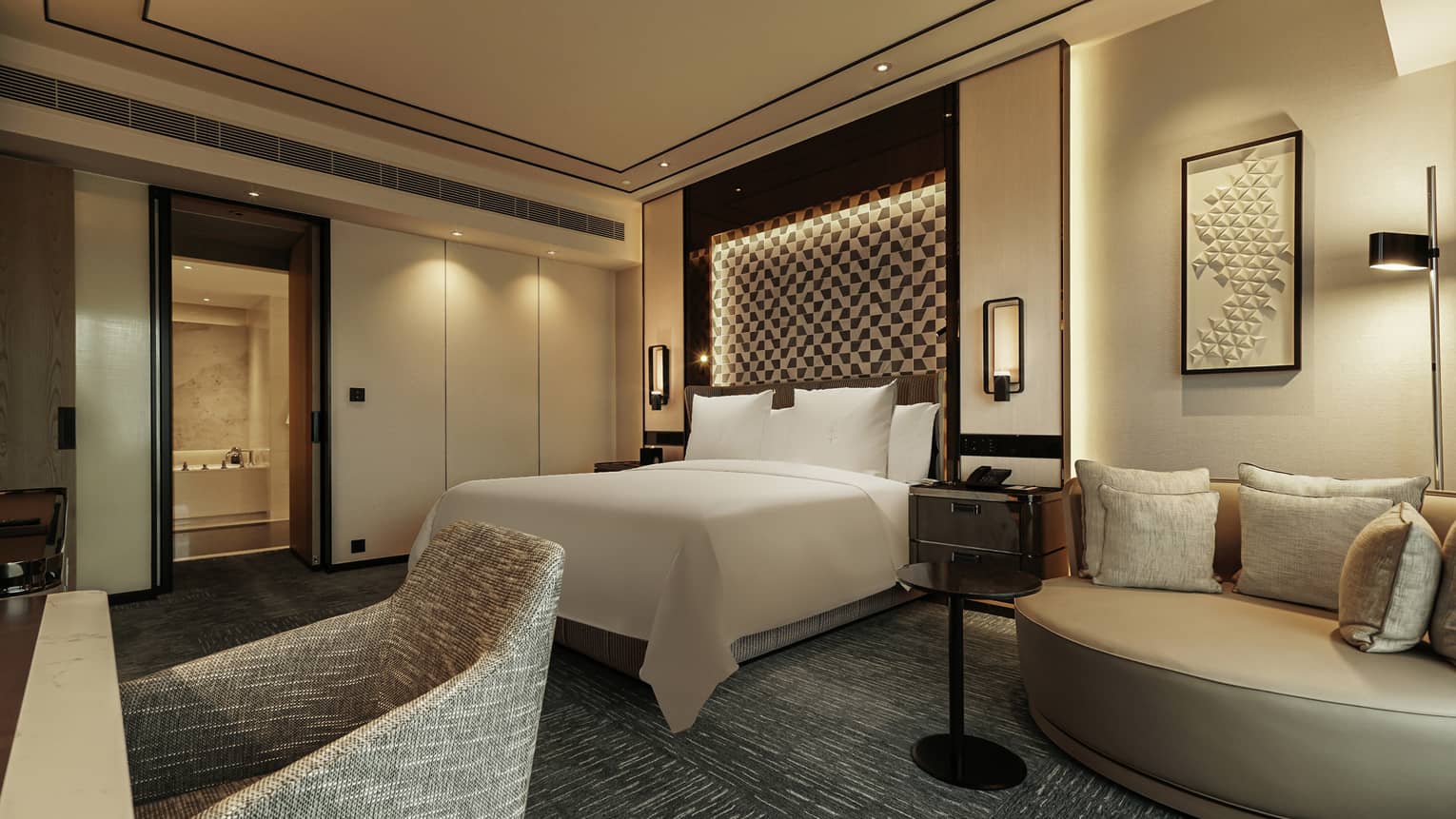 Luxurious hotel bedroom with large bed, modern geometric headboard, accent lighting and seating area with cushions.