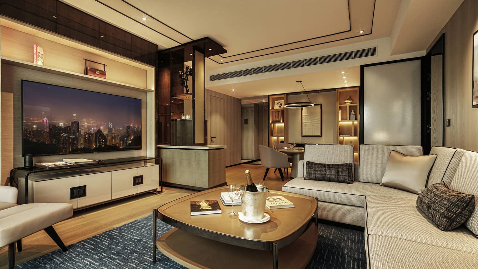 Luxurious hotel living room with beige sectional sofa, large TV displaying city skyline, coffee table with champagne and dining area in background.