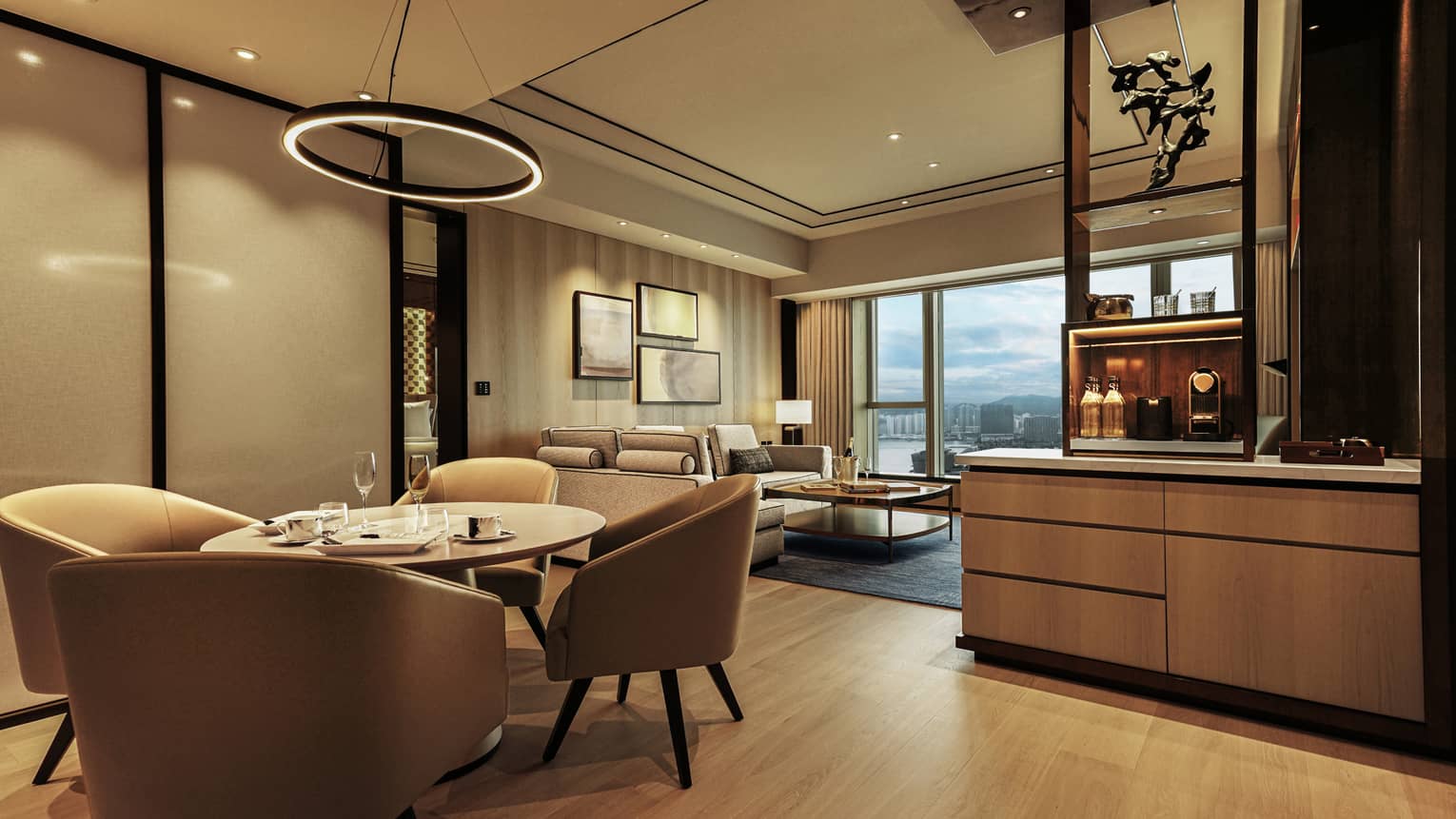Elegant hotel suite with dining area, beige chairs, round table, modern living room, wall art, city view window and coffee station.