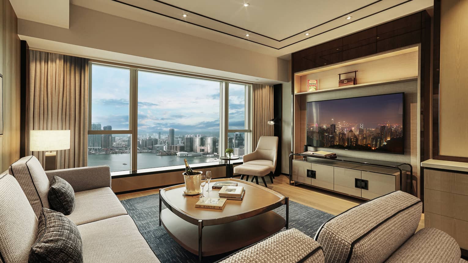Modern hotel living room with city and harbor view, beige sofas, armchair, TV displaying night skyline and coffee table with champagne.