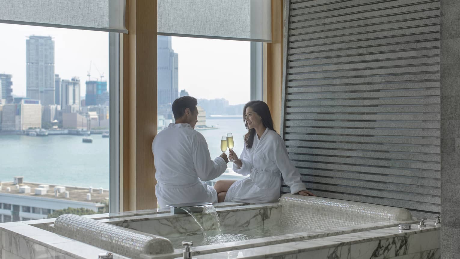 Man and woman clinking champagne glasses next to a private hot tub overlooking city harbour