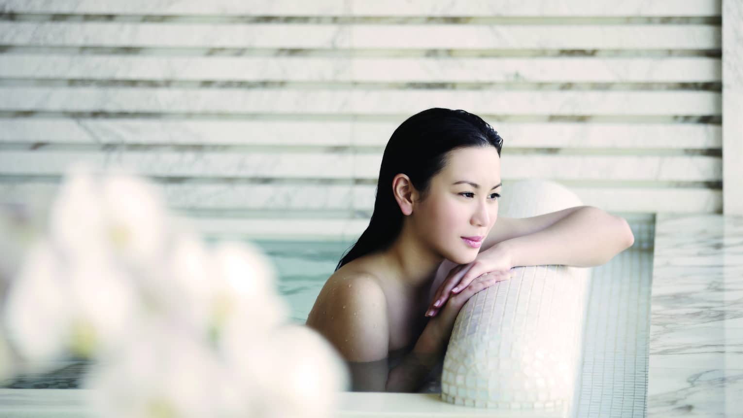 Woman with long wet hair rests arms, chin on white tile ledge of spa tub