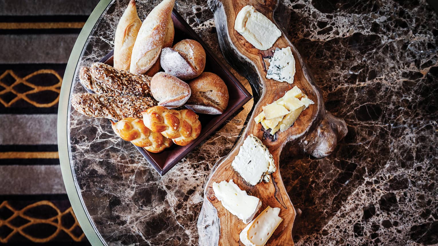 Assorted breads in a basket and a selection of cheeses on a rustic wooden board, set on a marble table.