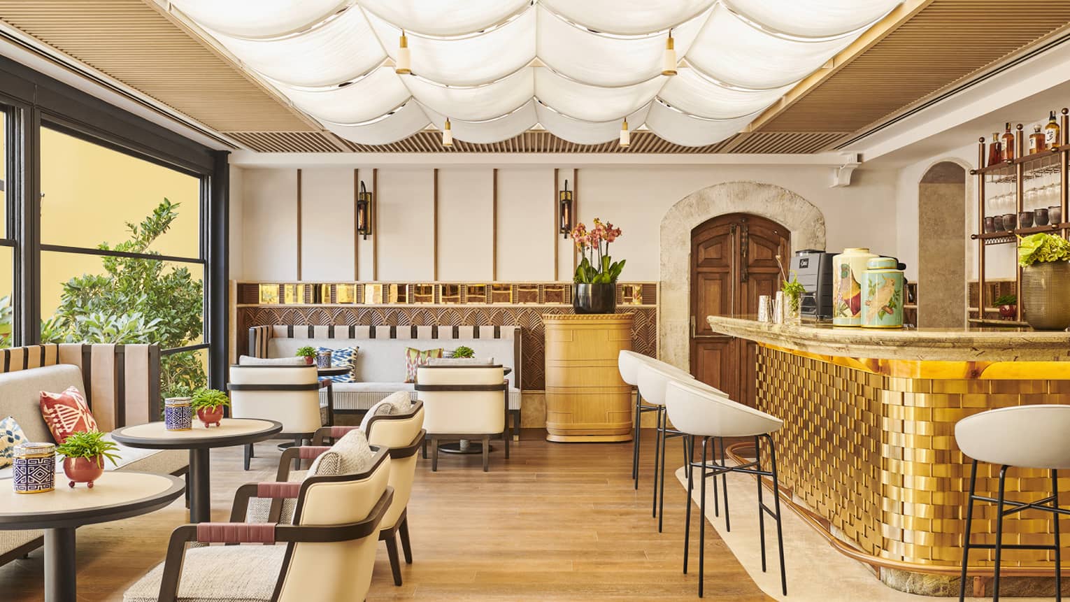 A well lit dining outlet with white chairs, large windows and a gold bar.