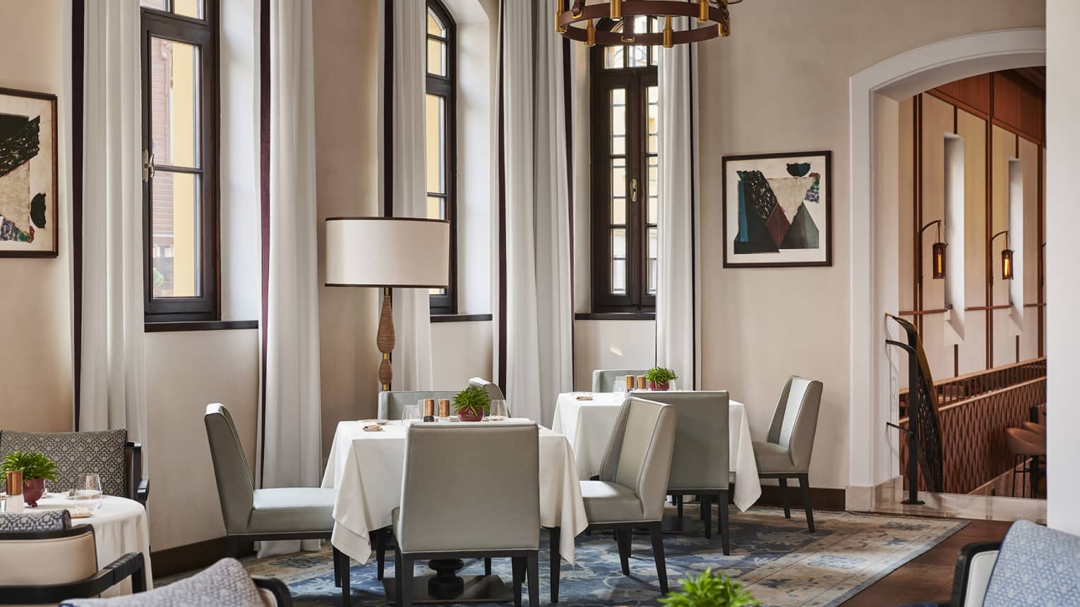 A dining outlet with square white cloth tables, arched windows and rugs on hard floor.
