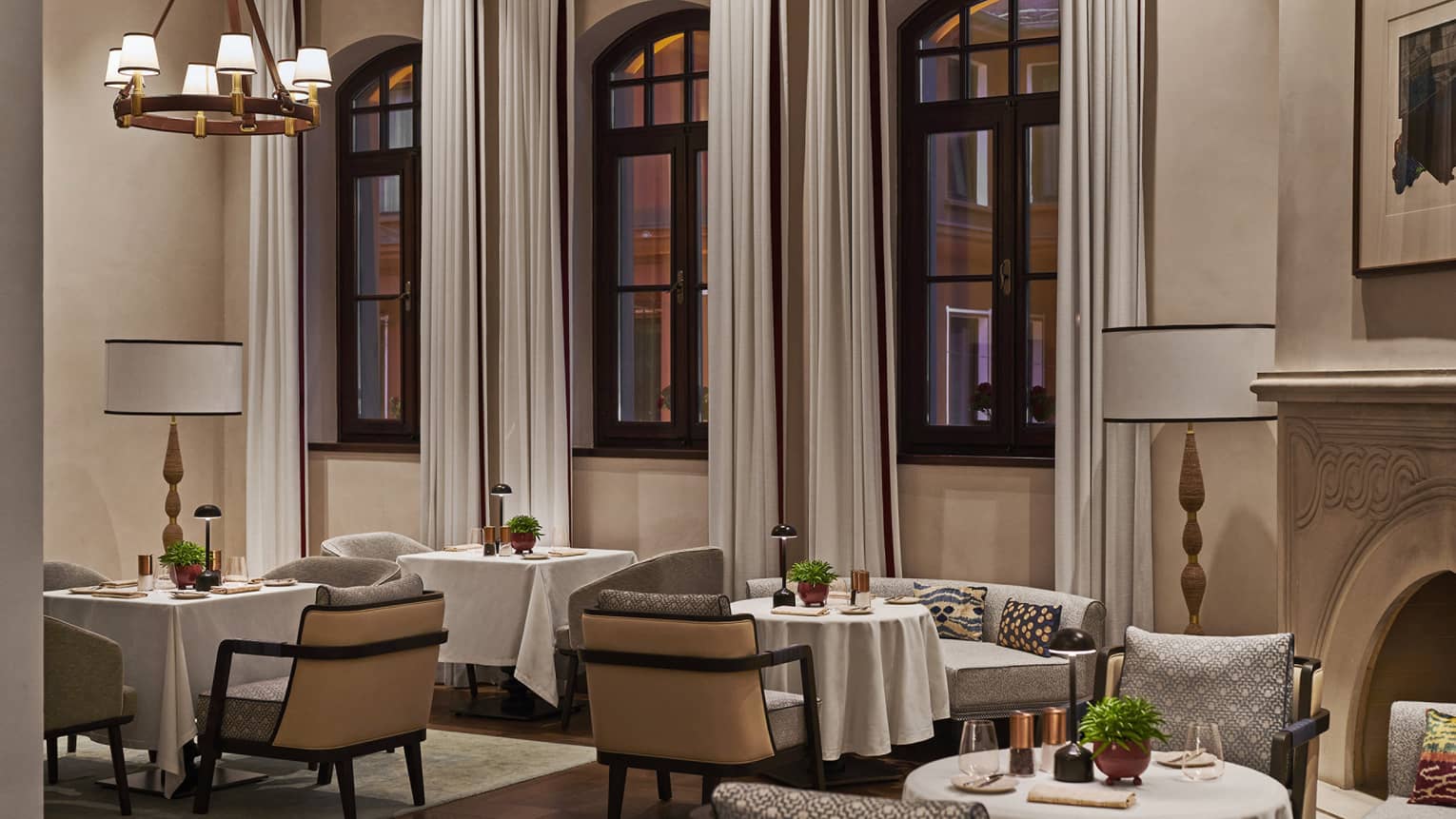 A well lit dining area with white cloth tables, chairs with pillows and large arched windows.