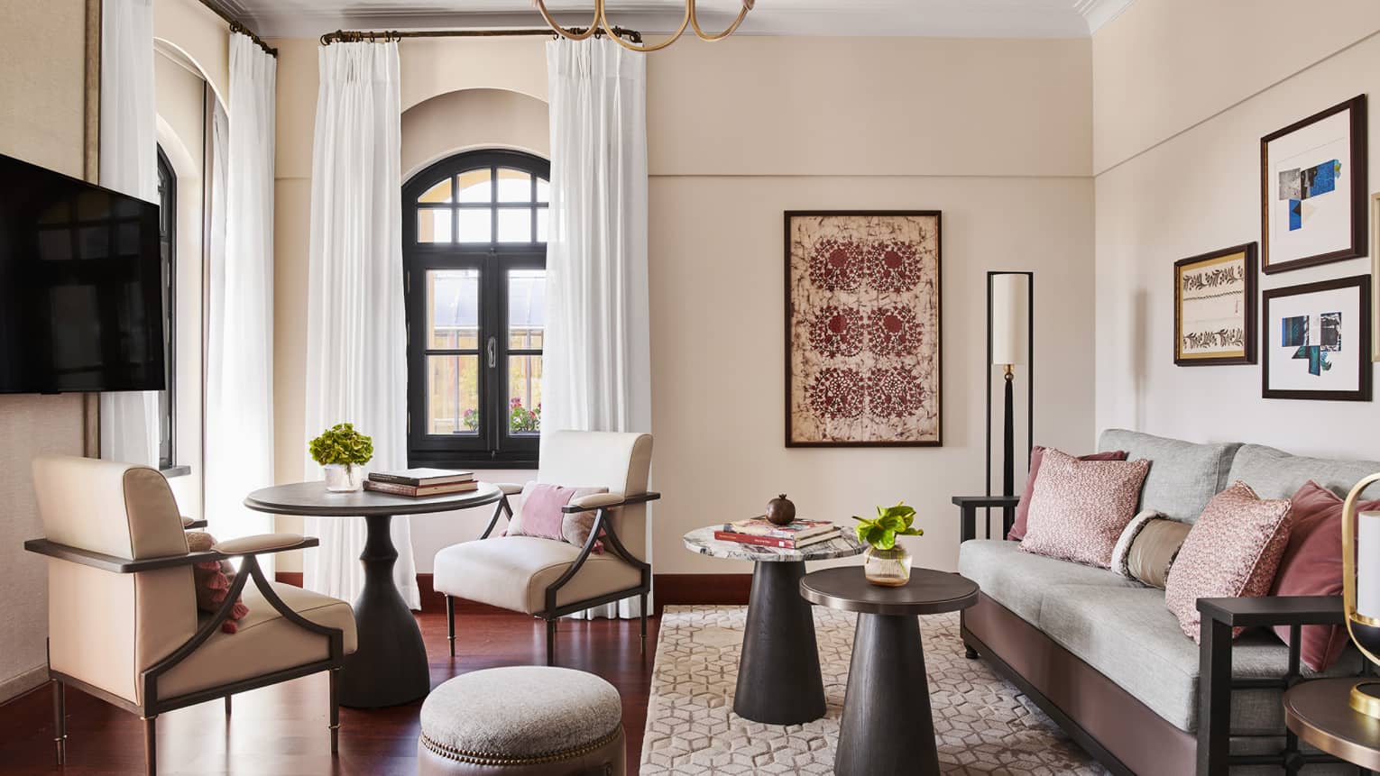 Living room with sofa, two small wooden tables, stool, round dining table with two chairs, TV and two arched windows