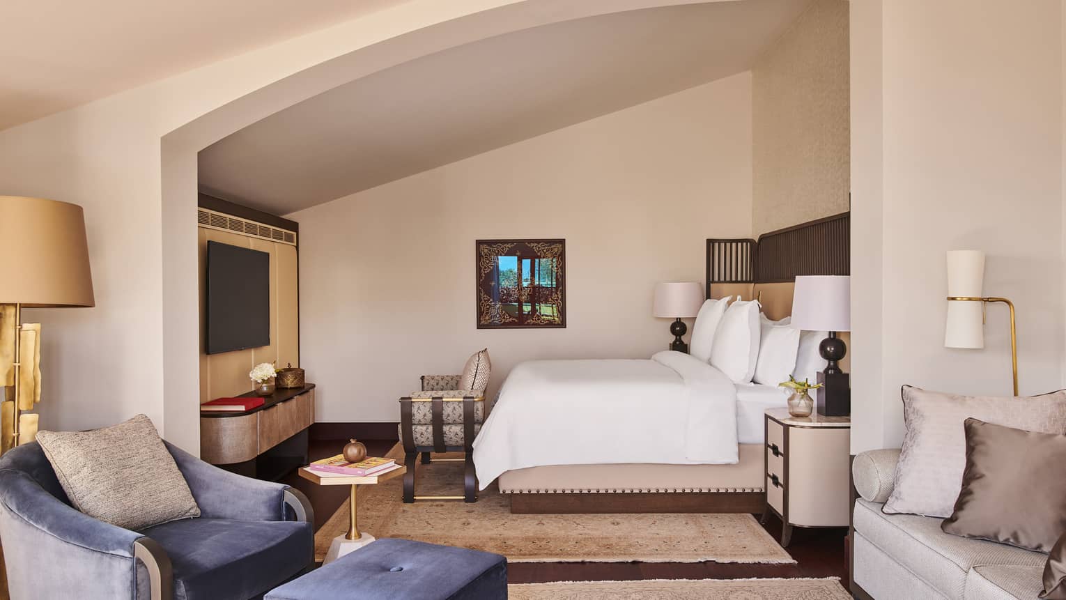 View through hotel suite’s living room to white Four Seasons bed tucked into angled alcove with chair at food and flat-screen TV on wall