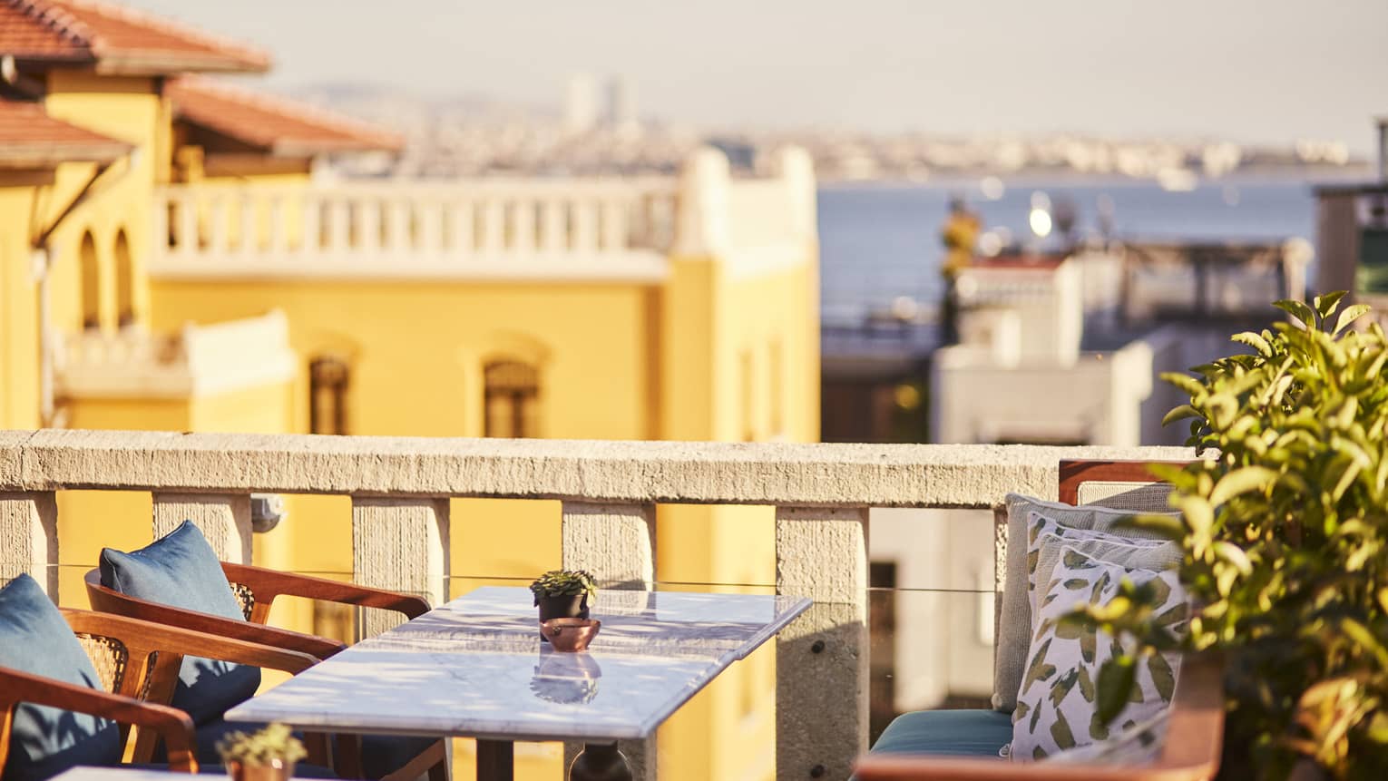 Marble table flanked by wood chairs and bench on a rooftop patio, views of beige and yellow buildings, the sea beyond. 
