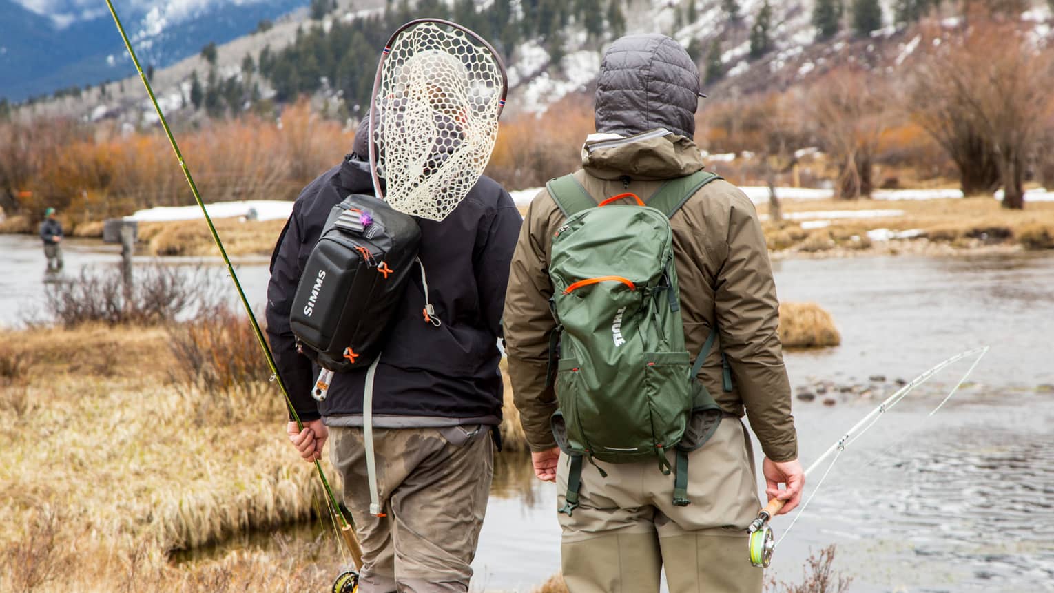 Two people with backpacks and fishing rods look at river with snowy mountain in background 