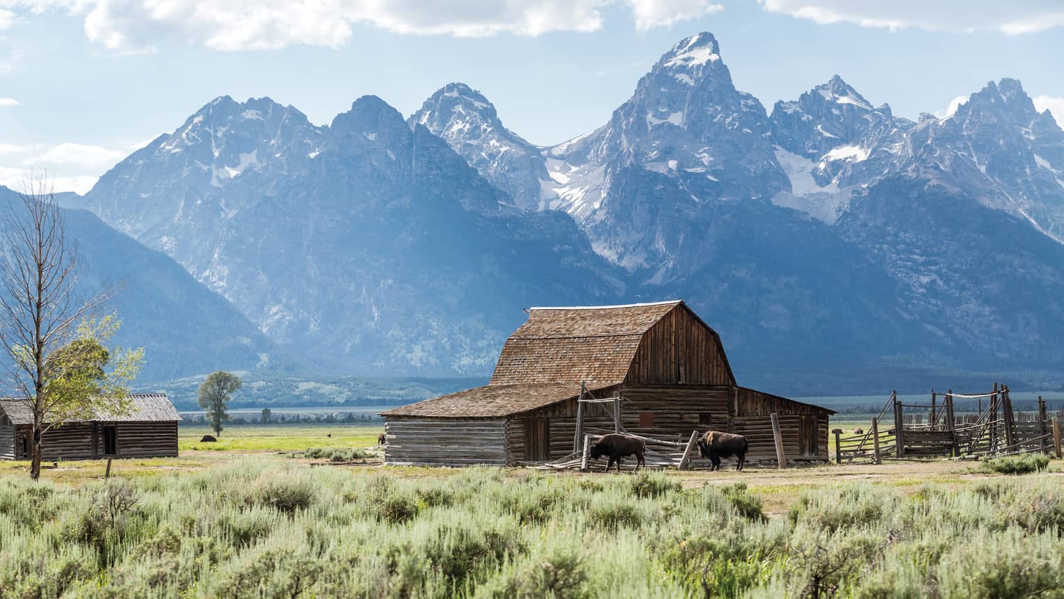 A wood barn in a green field under a partly cloudy sky, with trees in the foreground and dramatic mountains in the background.