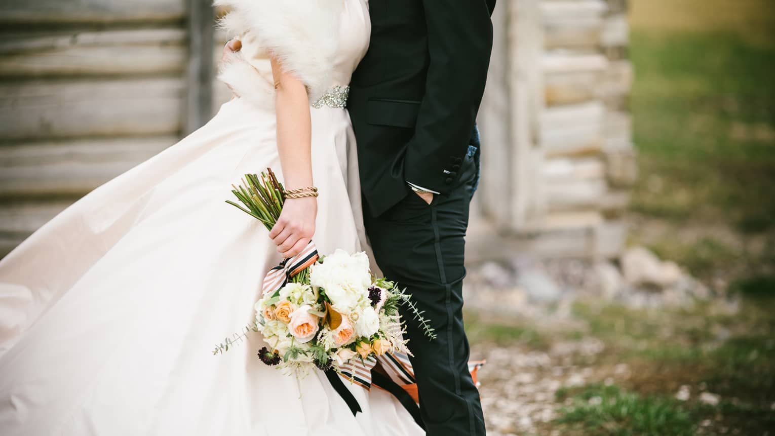 Groom embraces bride wearing fur shawl and holding long wedding floral bouquet