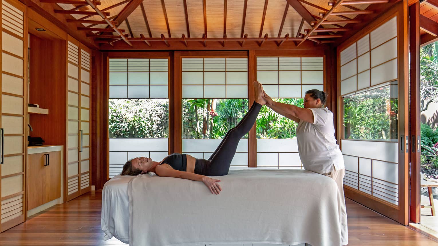 Wellness practitioner lifts legs of woman on spa treatment bed, in a private hale with tropical surroundings