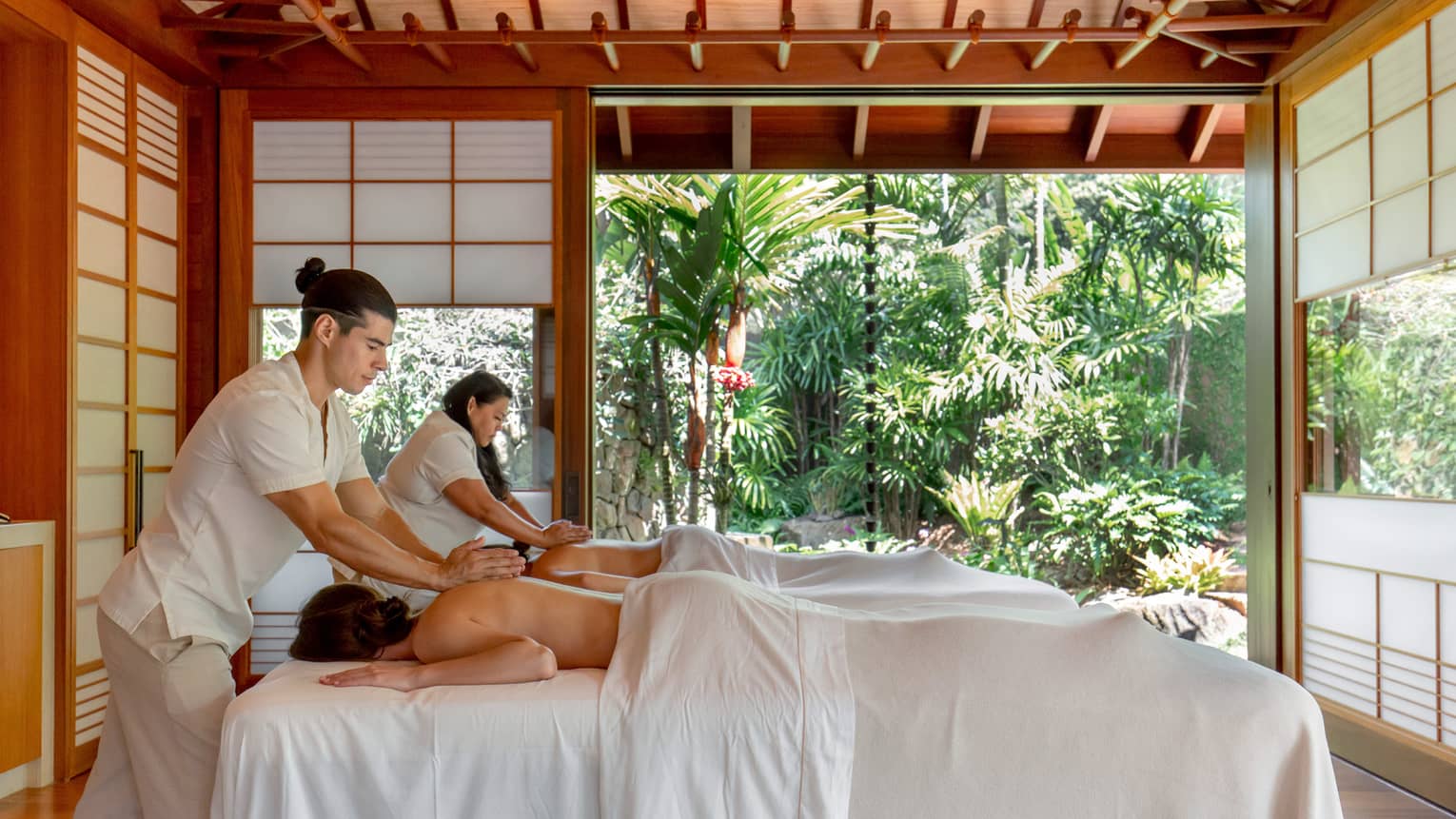 Two guests receive massages in a bright wooden hut with frosted glass windows and an entrance that opens to abundant trees.