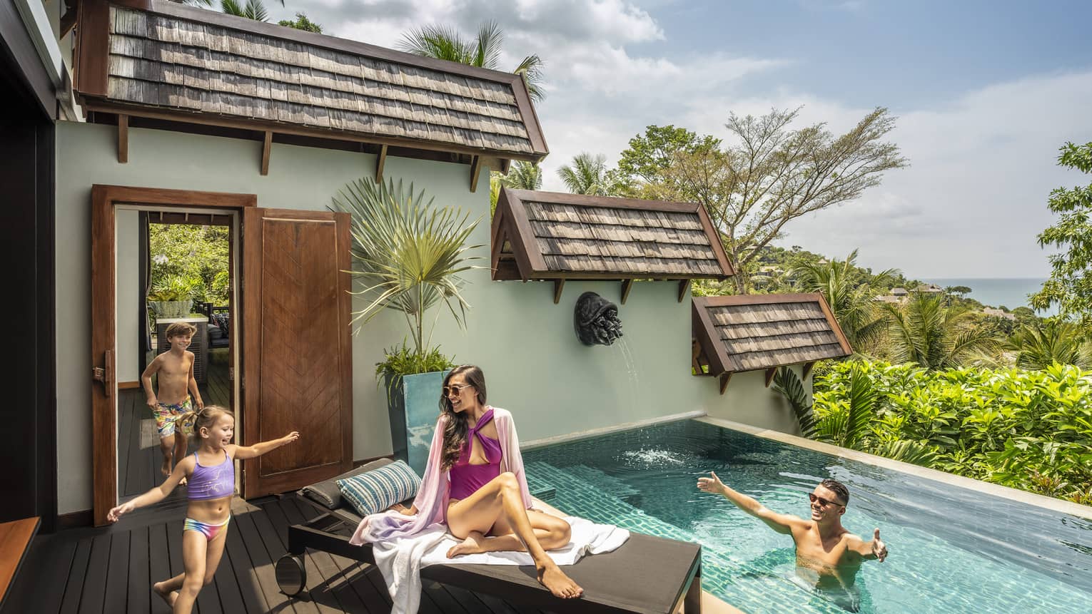 Two young children run into a small pool. Woman sits on lounge chair and man welcomes children in the pool.