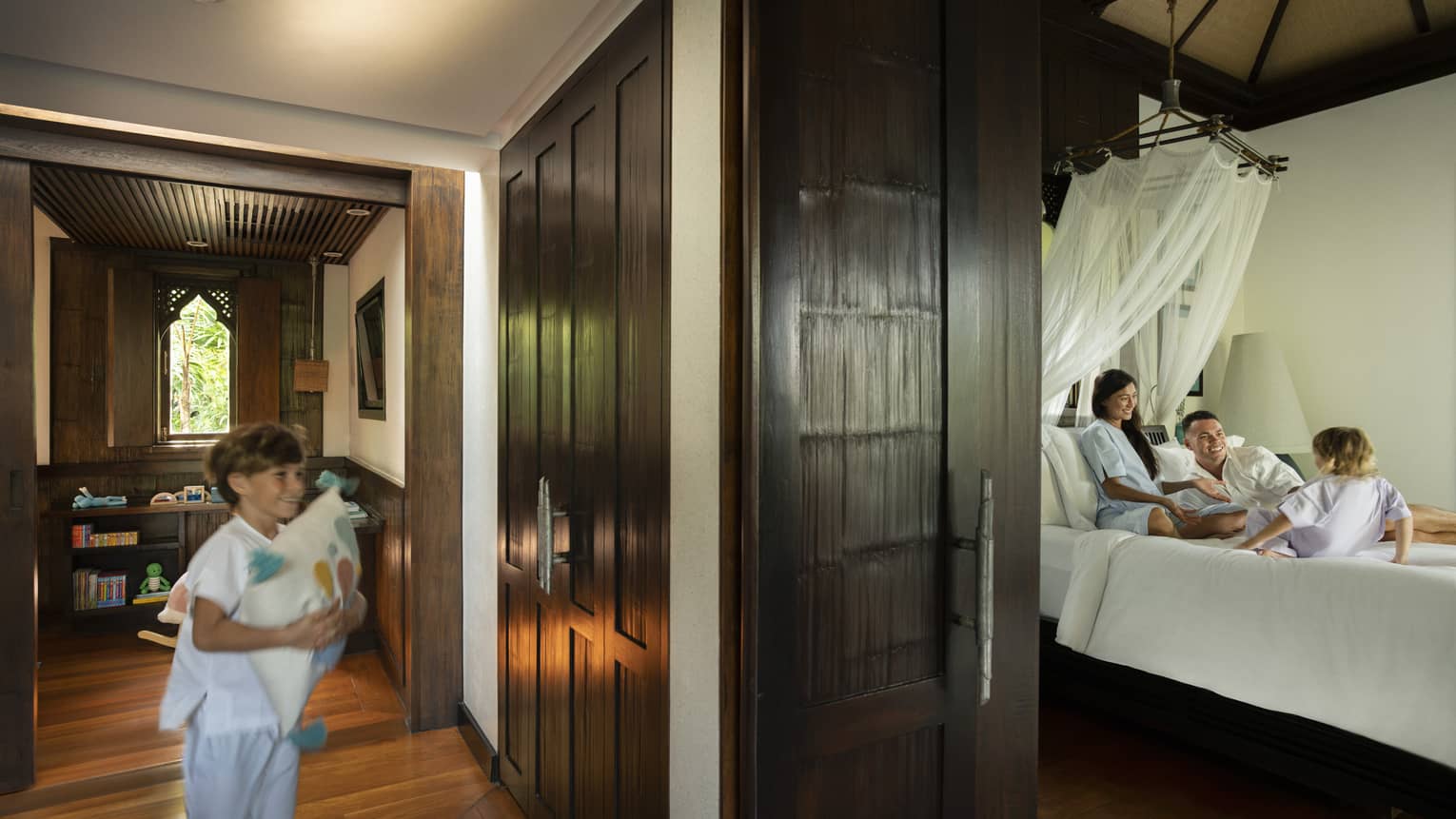 Young boy runs down a hallway to a bedroom where woman, man and small child play