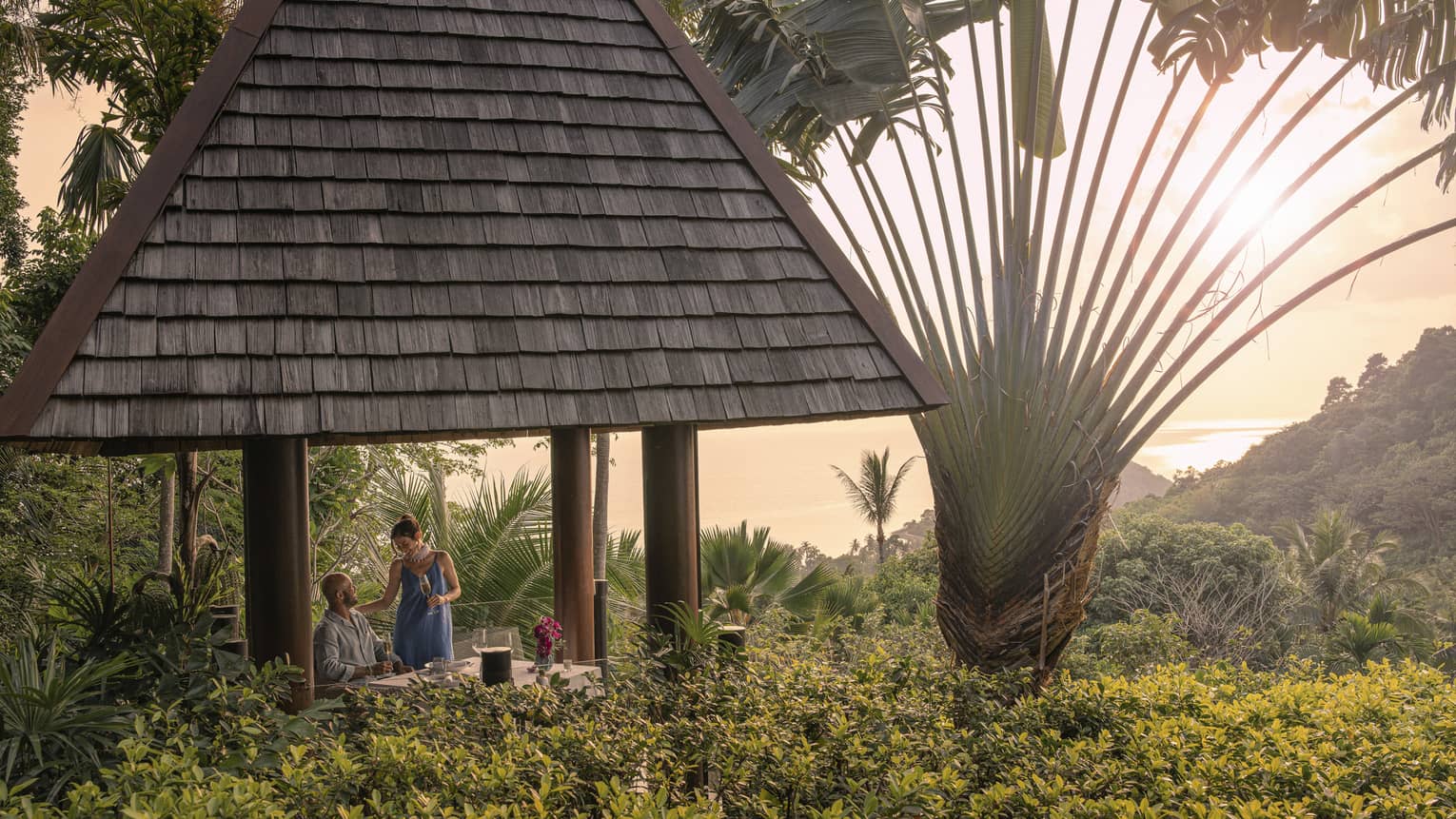 A couple with champagne in a tall hilltop pavilion with a dark, straw roof, set amidst lush forest and a giant fan palm tree.