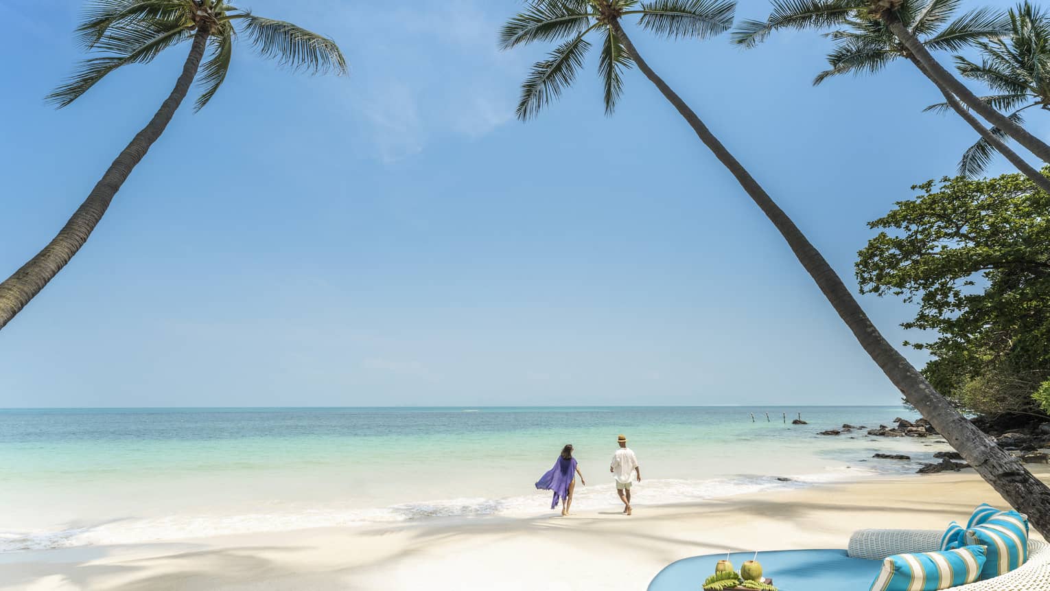Couple walks along the shoreline framed by palm trees, round turquoise lounge chair in the foreground