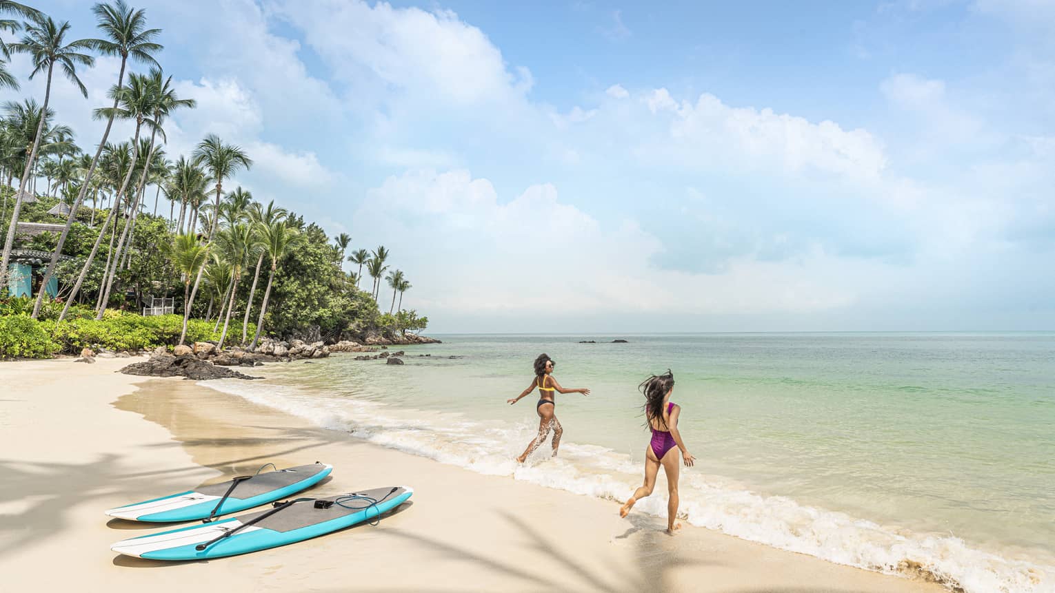 Two women in swimsuits wade into the water at the beach, their surfboards on the shore behind them