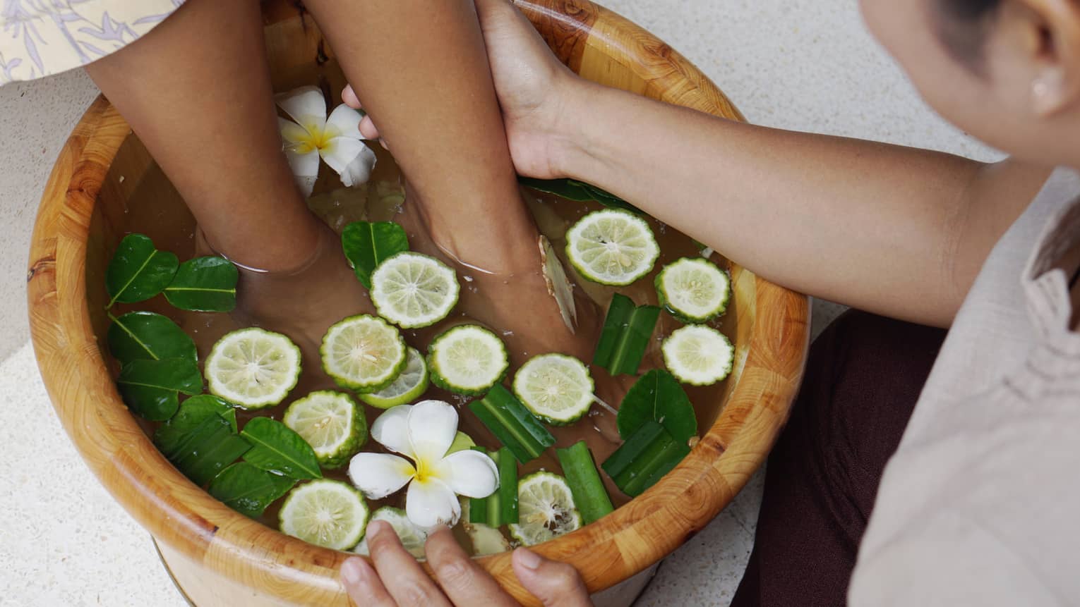 A guest receiving a foot massage at the resort spa in a wooden basin filled with water, lime slices, green leaves and white flowers.
