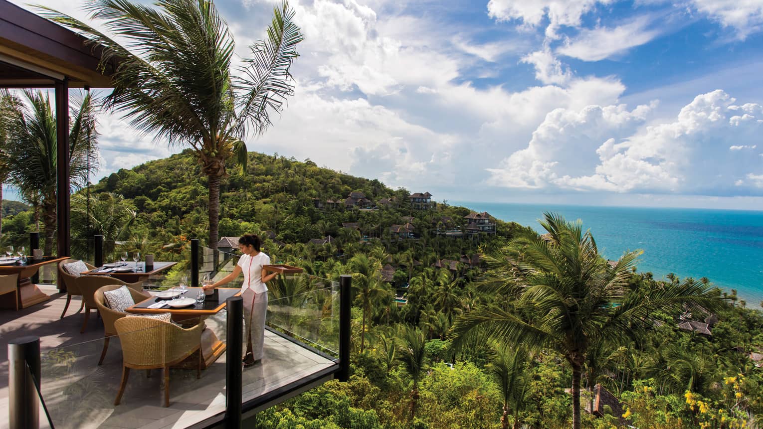 Server sets corner patio table behind glass balcony, high on tropical mountain