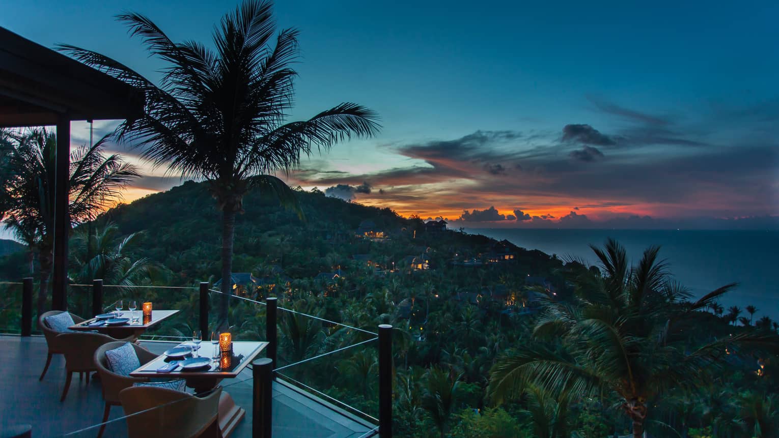 Corner of extended balcony with dining tables and chairs at dusk, expanse of forest, palm trees